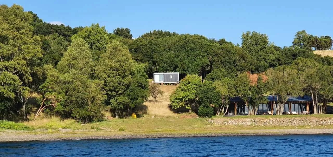 Vista del lago con cabañas y árboles a la orilla del agua, en un día soleado.