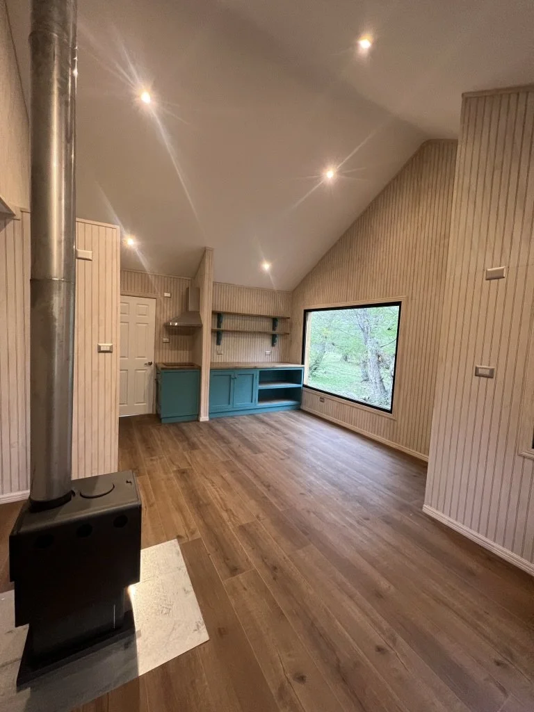 Interior de una habitación con paredes de madera clara, piso de madera, ventanas grandes y una estufa de leña negra.