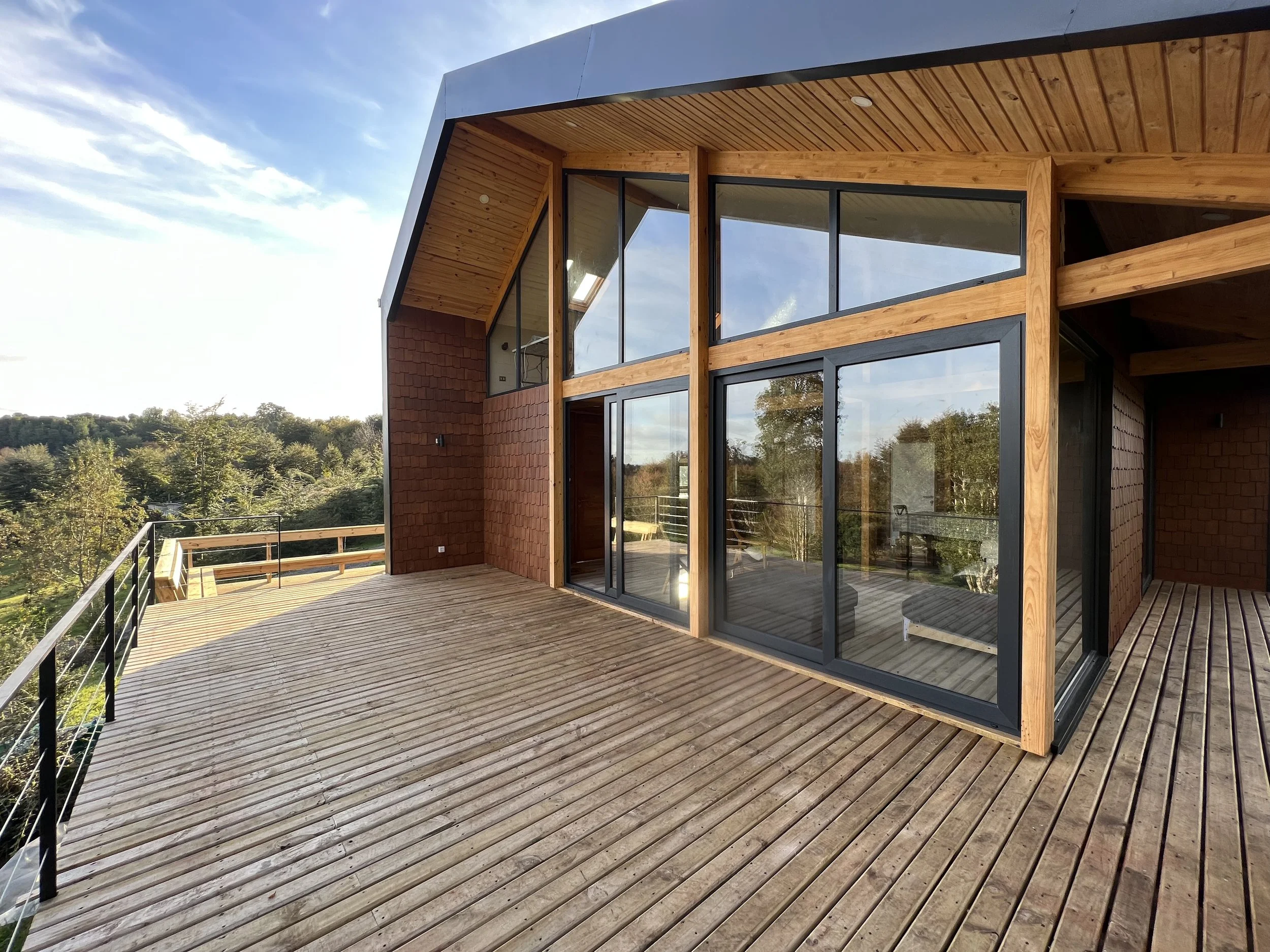 Terraza de madera en la esquina de una casa moderna con ventanas grandes de vidrio, rodeada de naturaleza y árboles en el fondo