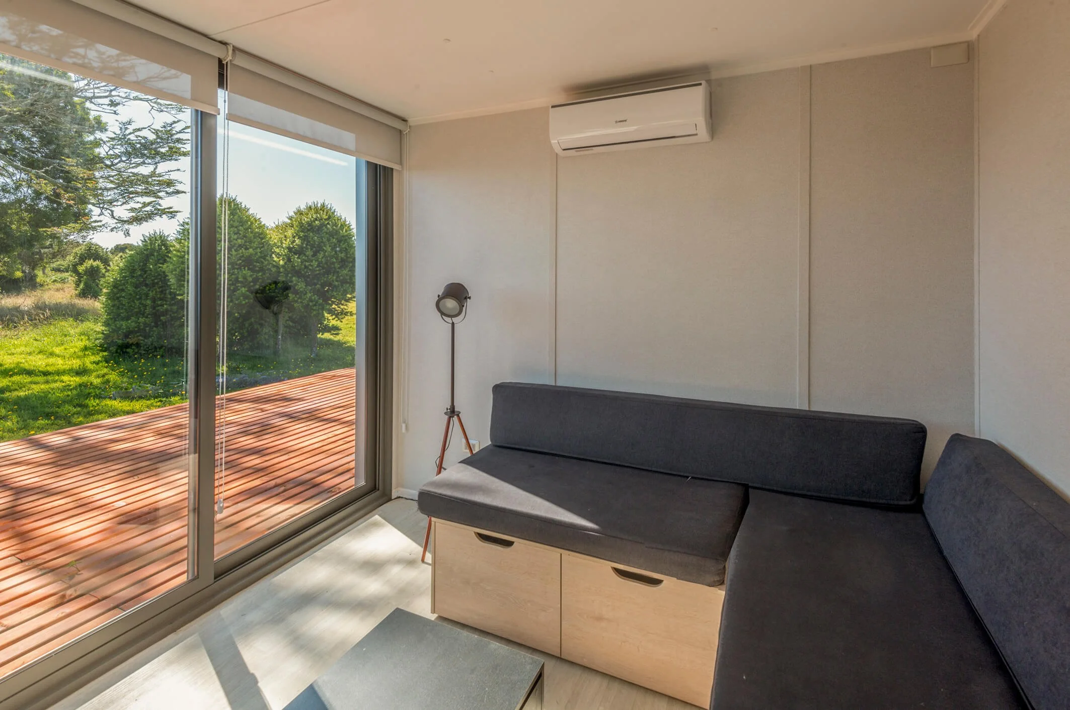 Interior de una sala con un sillón gris, una lámpara de pie y un aire acondicionado en la pared, con una vista de un jardín a través de una ventana grande.