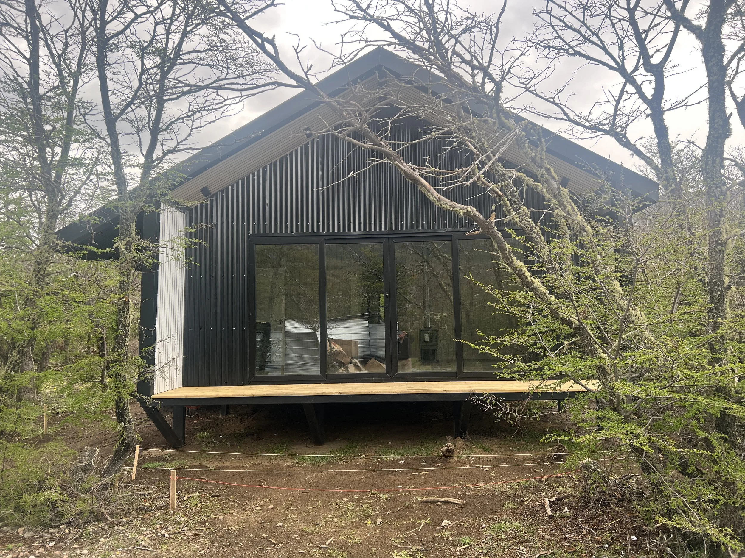 Casa moderna con paredes de metal negras y ventanas grandes, elevada sobre pilotes, en medio de árboles con ramas desnudas y algunas verdes, en un entorno boscoso.