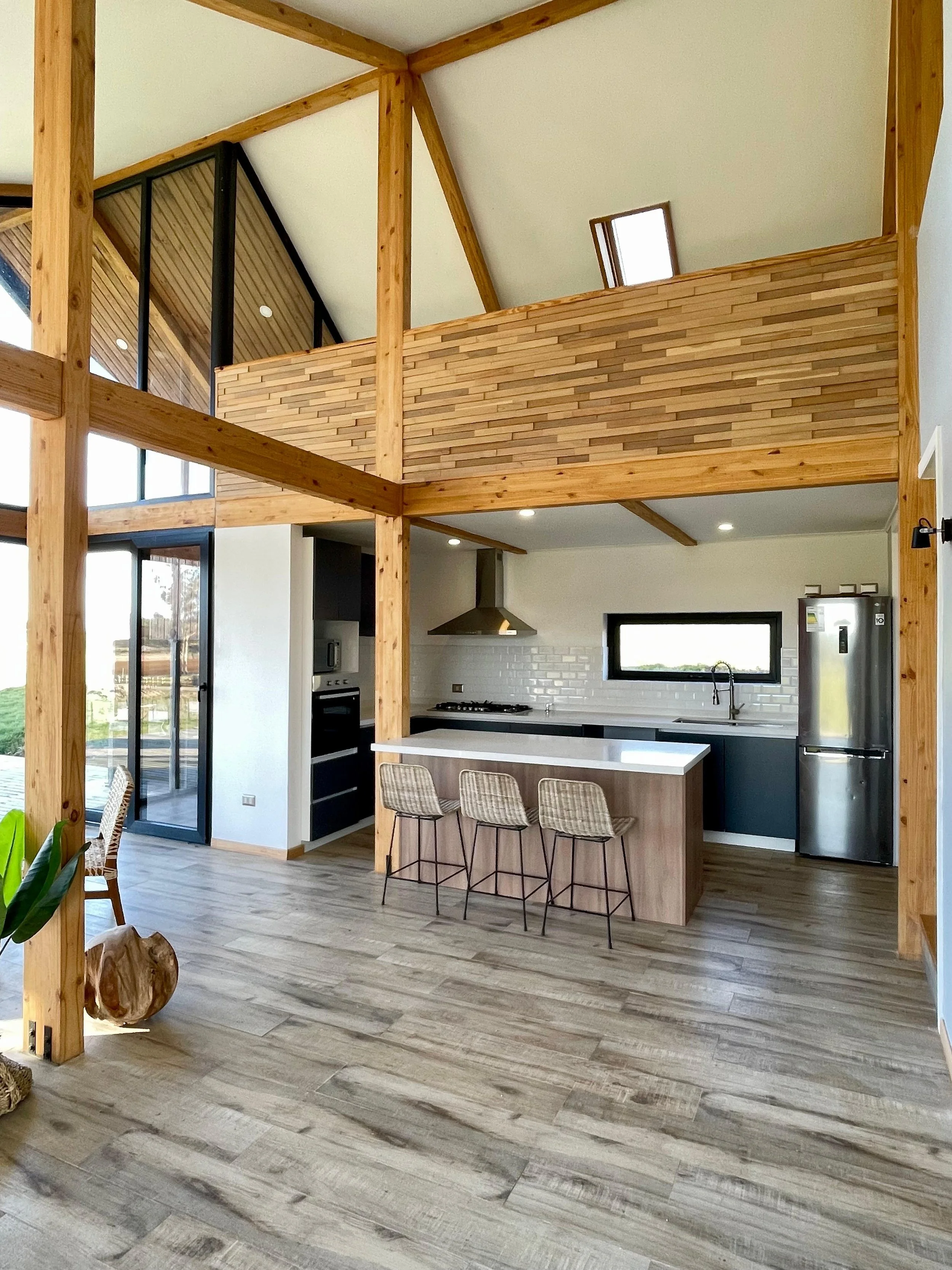 Interior de una sala moderna con estructura de madera, cocina con barra y sillas, y un balcón en la parte superior con paredes de ladrillos de madera.