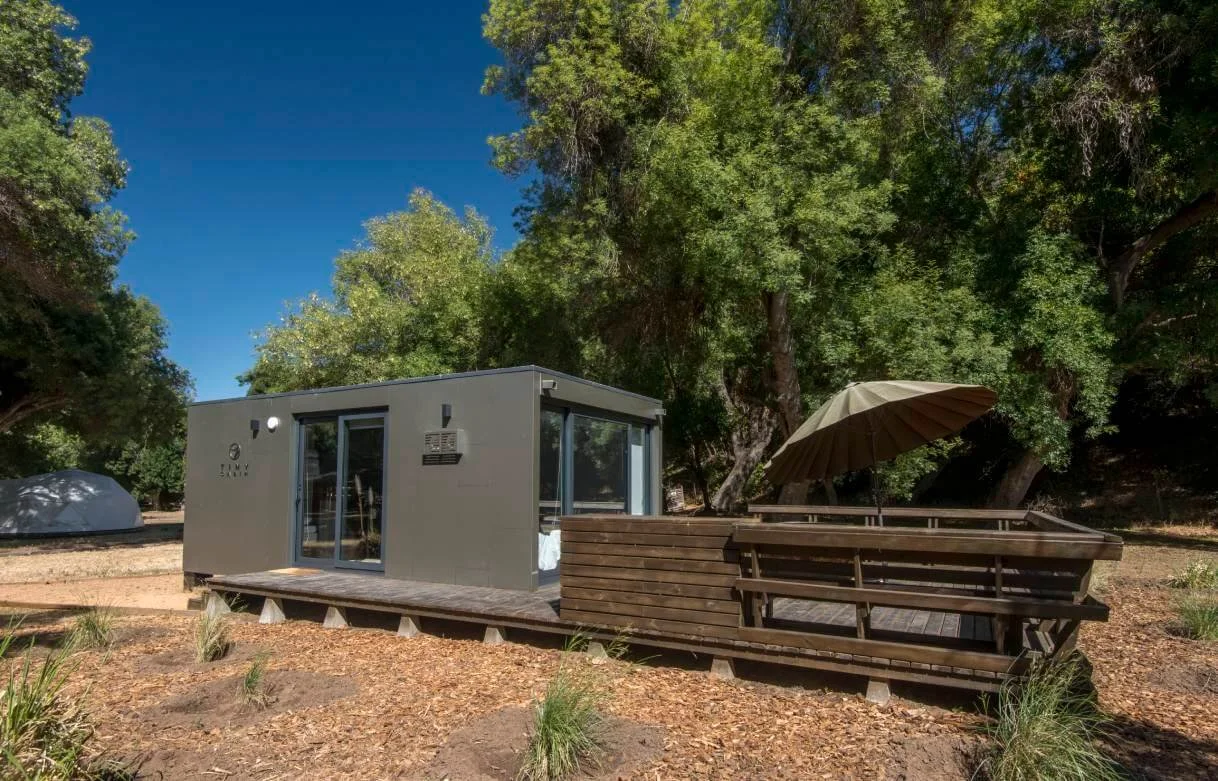 Casa moderna en un entorno natural rodeada de árboles, con una estructura de madera, un paraguas y áreas de descanso al aire libre.