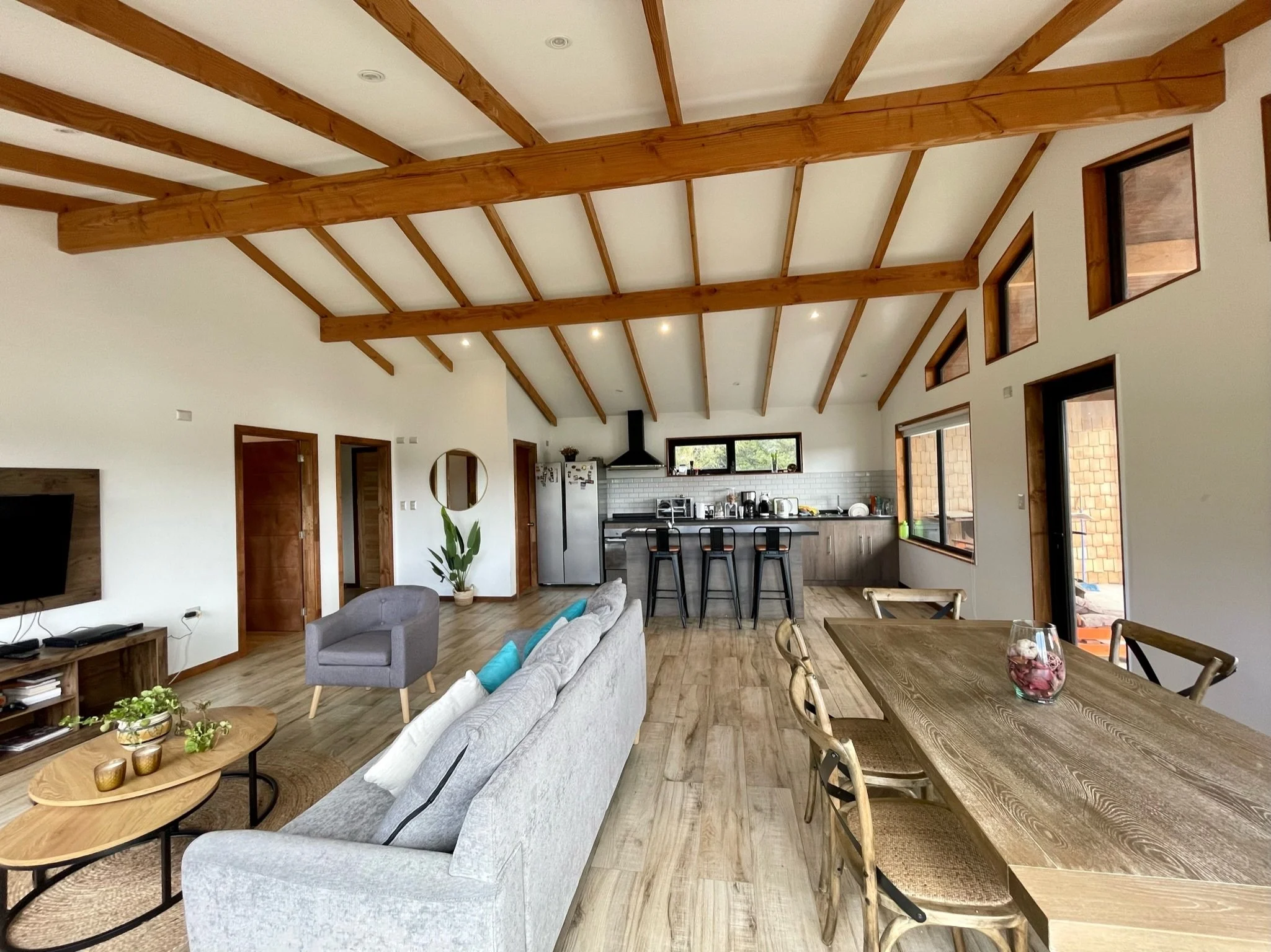 Interior de una sala de estilo rústico con vigas de madera en el techo, área de sala con sofá gris, sillón y mesa de centro, área de comedor con mesa grande y sillas de madera, en la cocina hay un fregadero, refrigerador, estufa y barra con bancos. V