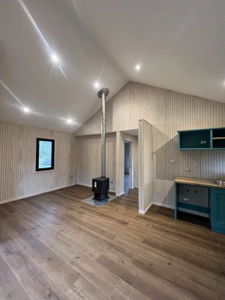 Sala vacía con paredes de madera, suelo de madera, una ventana, y una estufa de pellets en el centro, con una pequeña cocina azul.