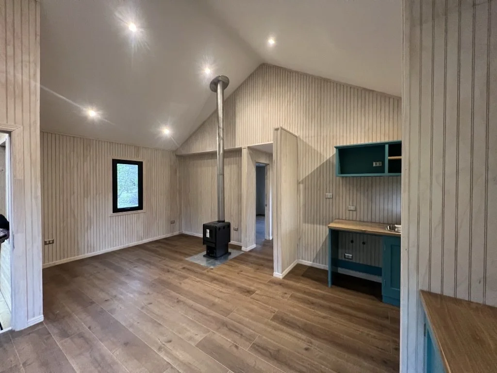 Sala con madera clara, paredes de madera a rayas, estufa de leña y pequeña cocina con gabinetes azules.