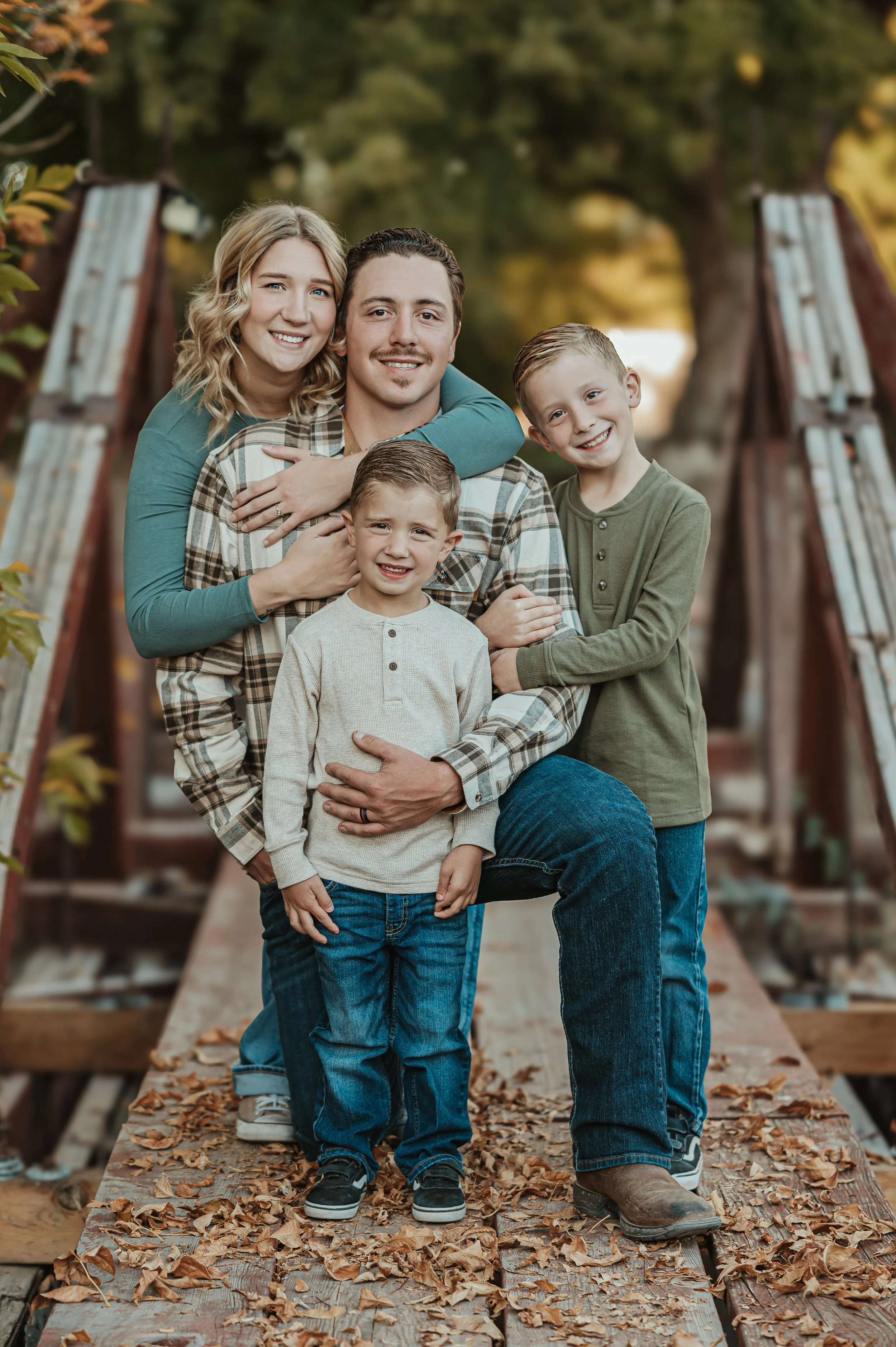 Family of four outdoors on a wooden bridge covered with fallen leaves, smiling and posing for a photo, with trees in the background.