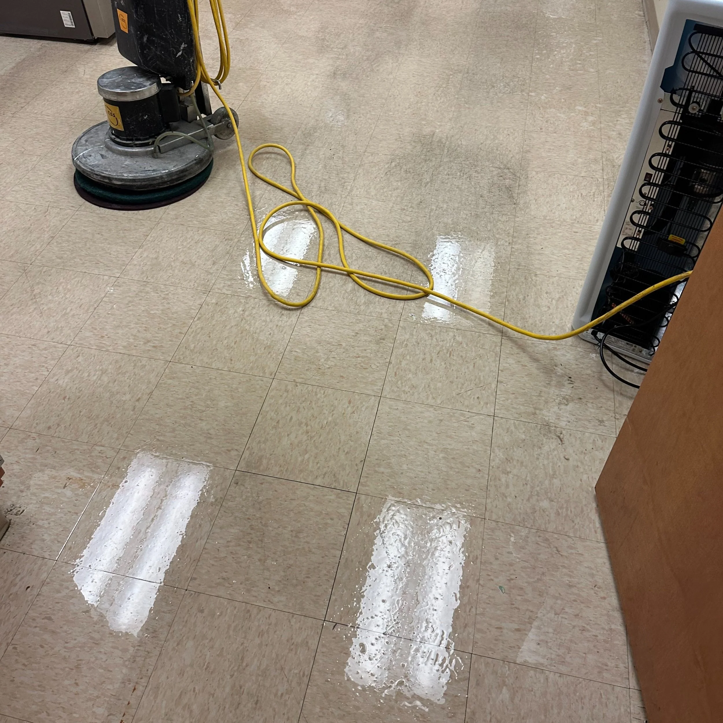 Floor buffer machine with yellow extension cord plugged in, cleaning a shiny, wet tiled floor in a commercial or retail space.