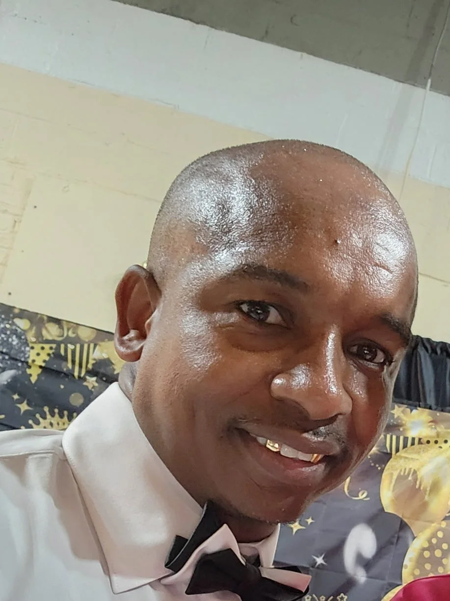 A man with a shaved head wearing a white dress shirt and black bow tie smiling at the camera in front of a decorated background.