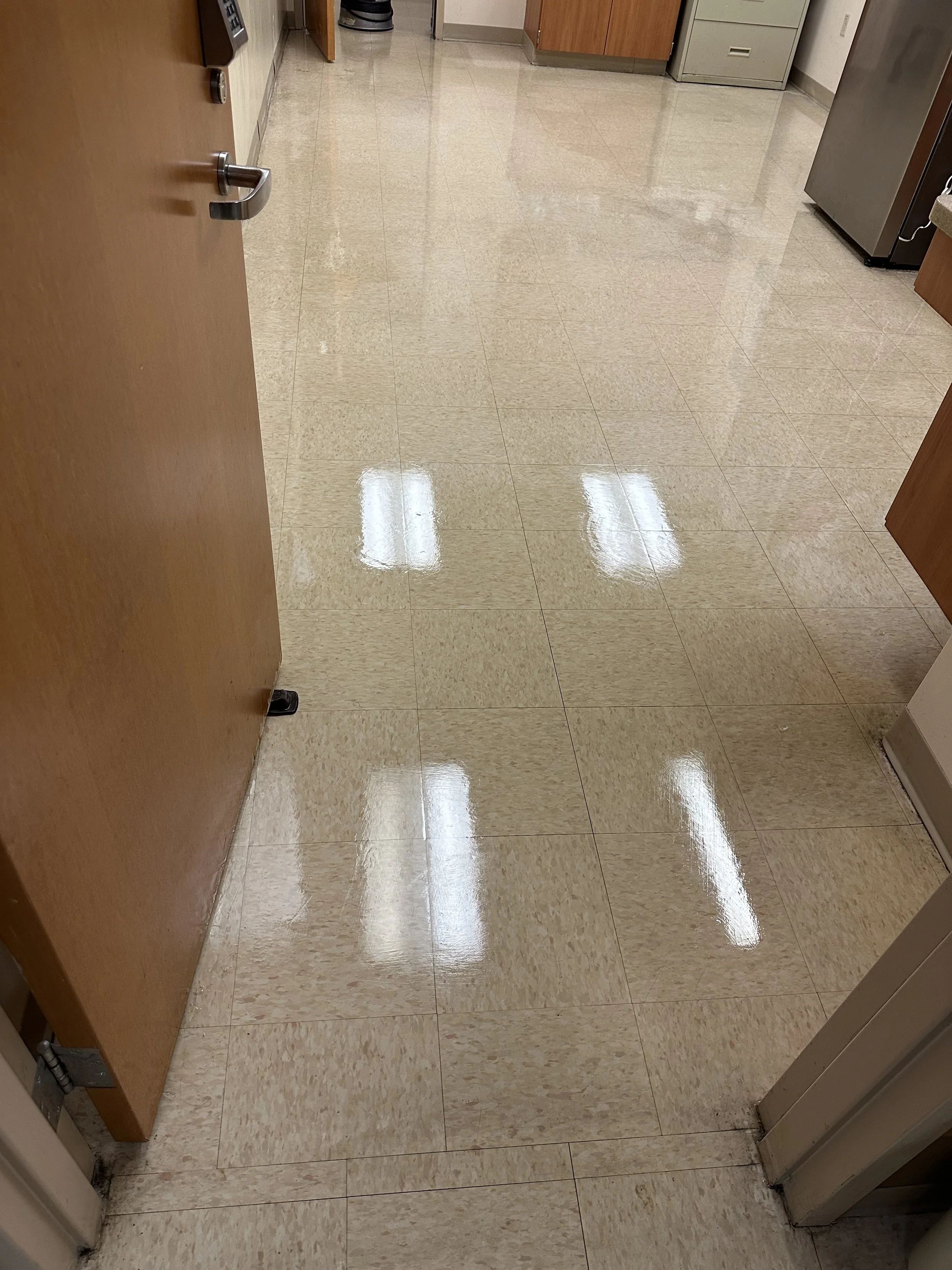 Clean, shiny beige tile floor in a room with wooden cabinets, a metal refrigerator, and a wooden door partially open.