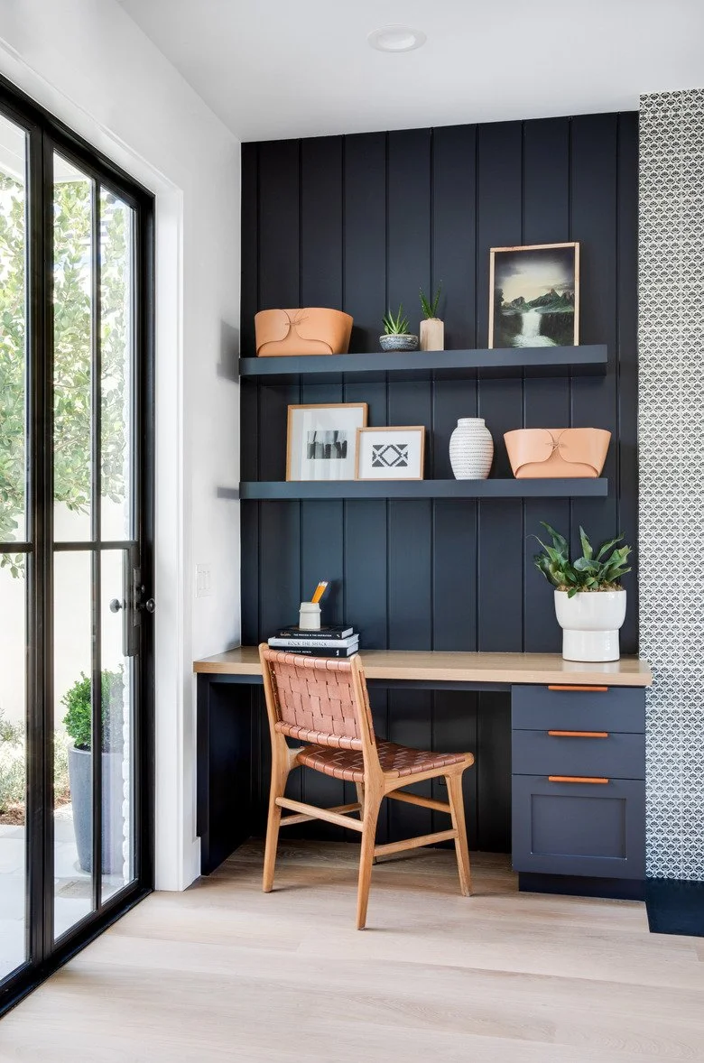 Home office corner with a built-in desk, navy blue paneled wall, floating shelves with decorative items, and a window with black framing on the left.