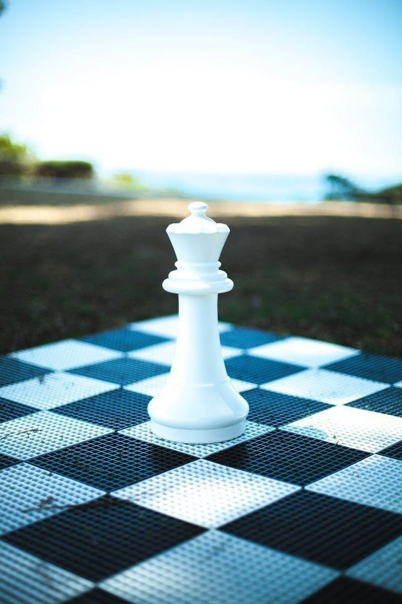 A large white chess king piece on a black and white checkered chessboard outdoors, with trees and a body of water in the background.