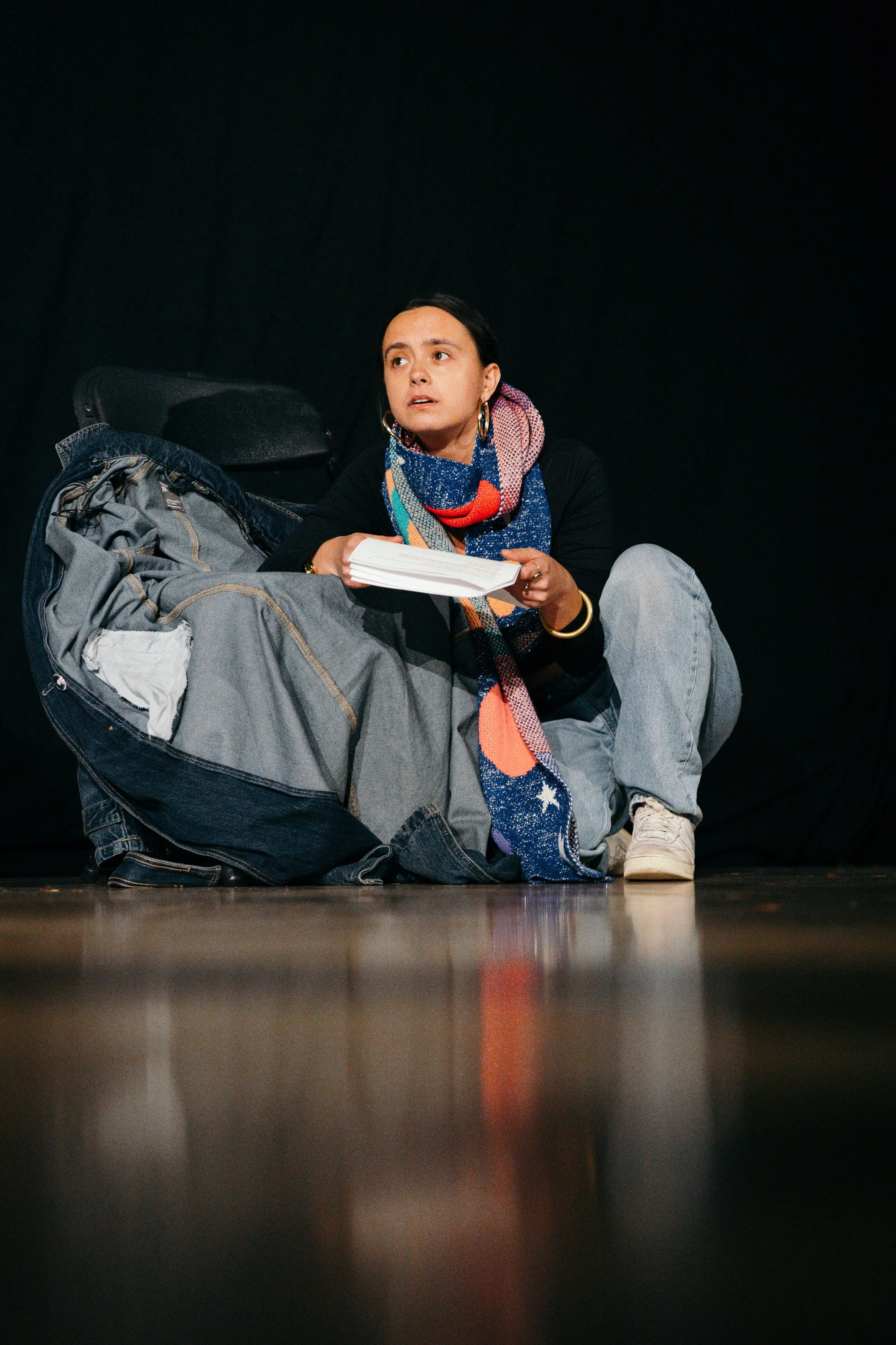 A woman sitting on the floor with a large bag, holding a book, and looking worried or concerned against a black background.