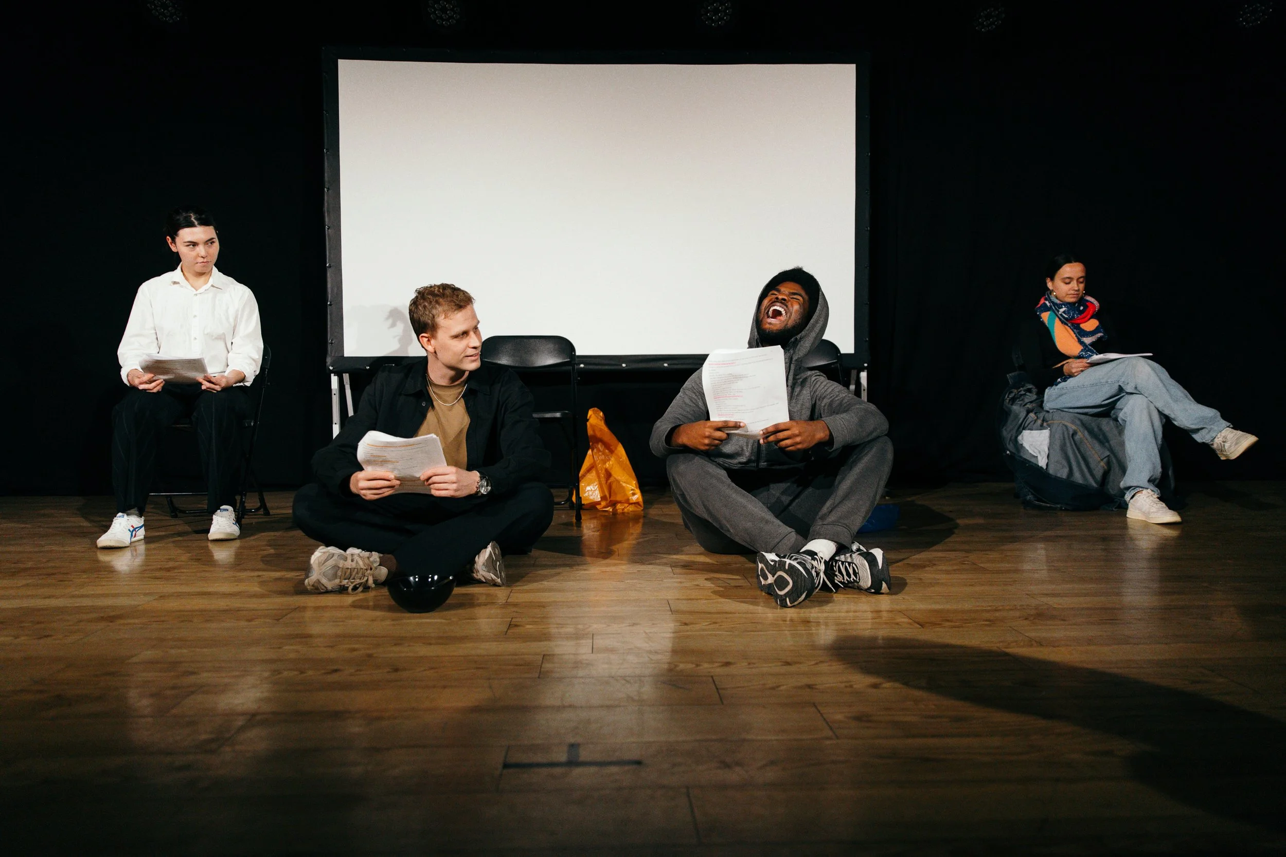 Four people sitting on a stage with a large white screen behind them. The person on the far left is a woman holding papers, the second person from the left is a man sitting cross-legged holding papers, the third person is a man sitting on the floor l