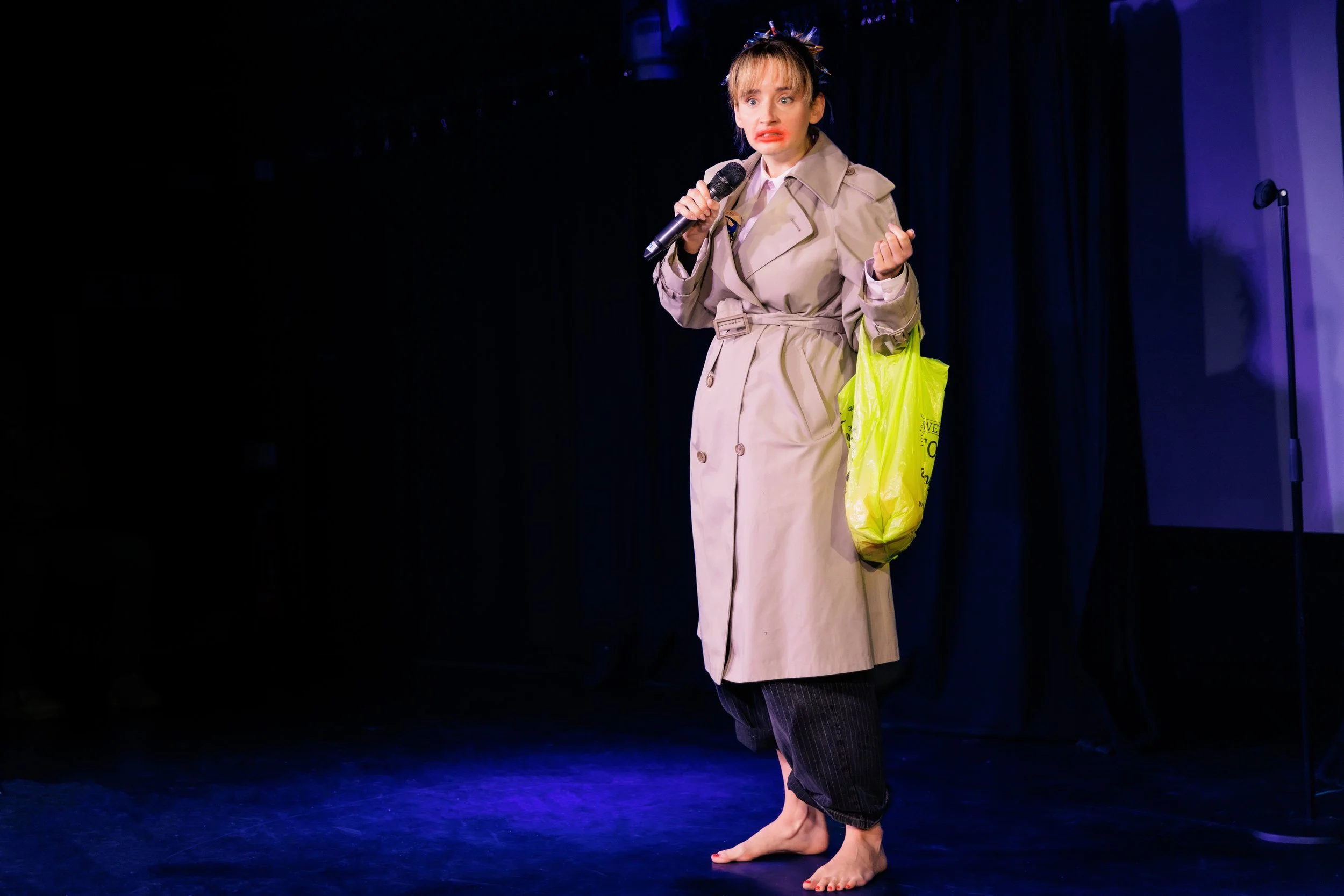 A woman performing on stage wearing a trench coat and striped pants, holding a microphone and a yellow shopping bag, with a dark curtain background.