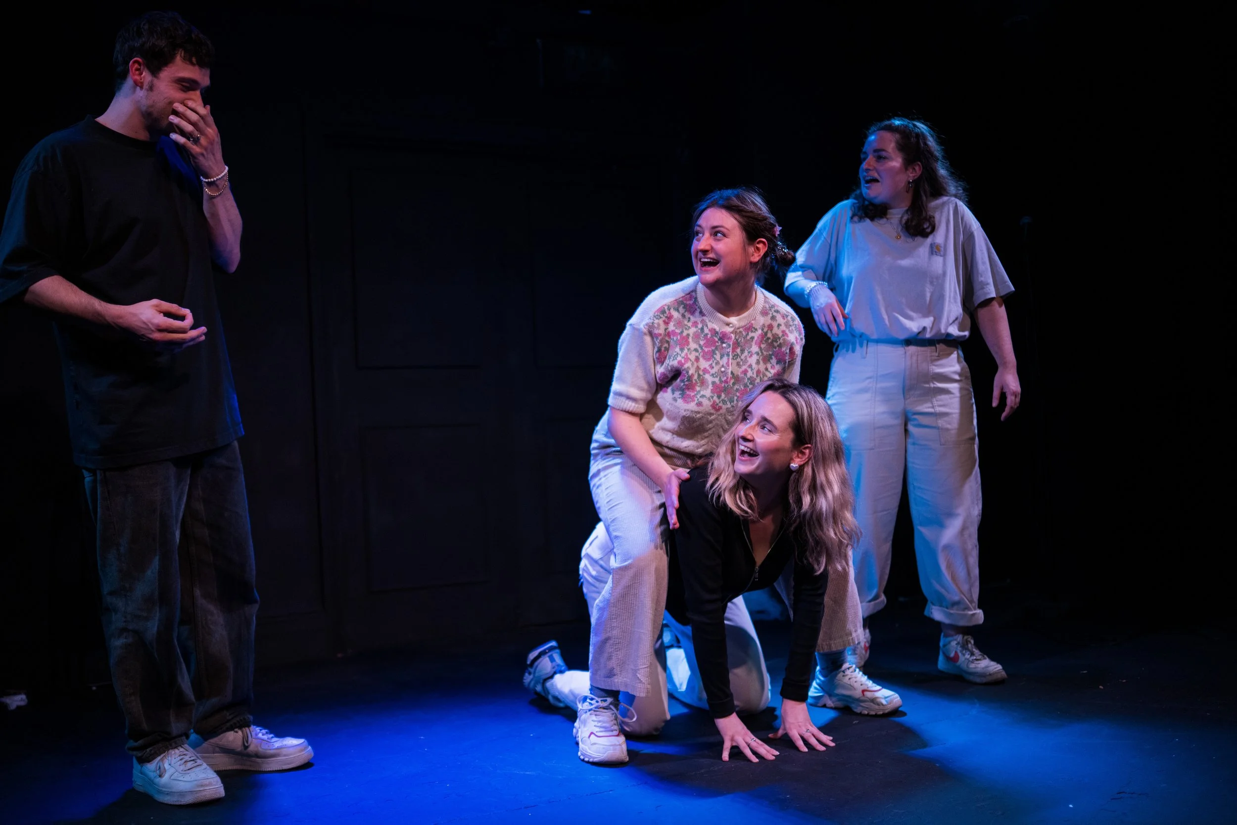 Four women and one man on stage, engaging in a theatrical performance under stage lighting. The man is on the left, covering his mouth, while three women are interacting with him and each other, all smiling and expressing amusement.