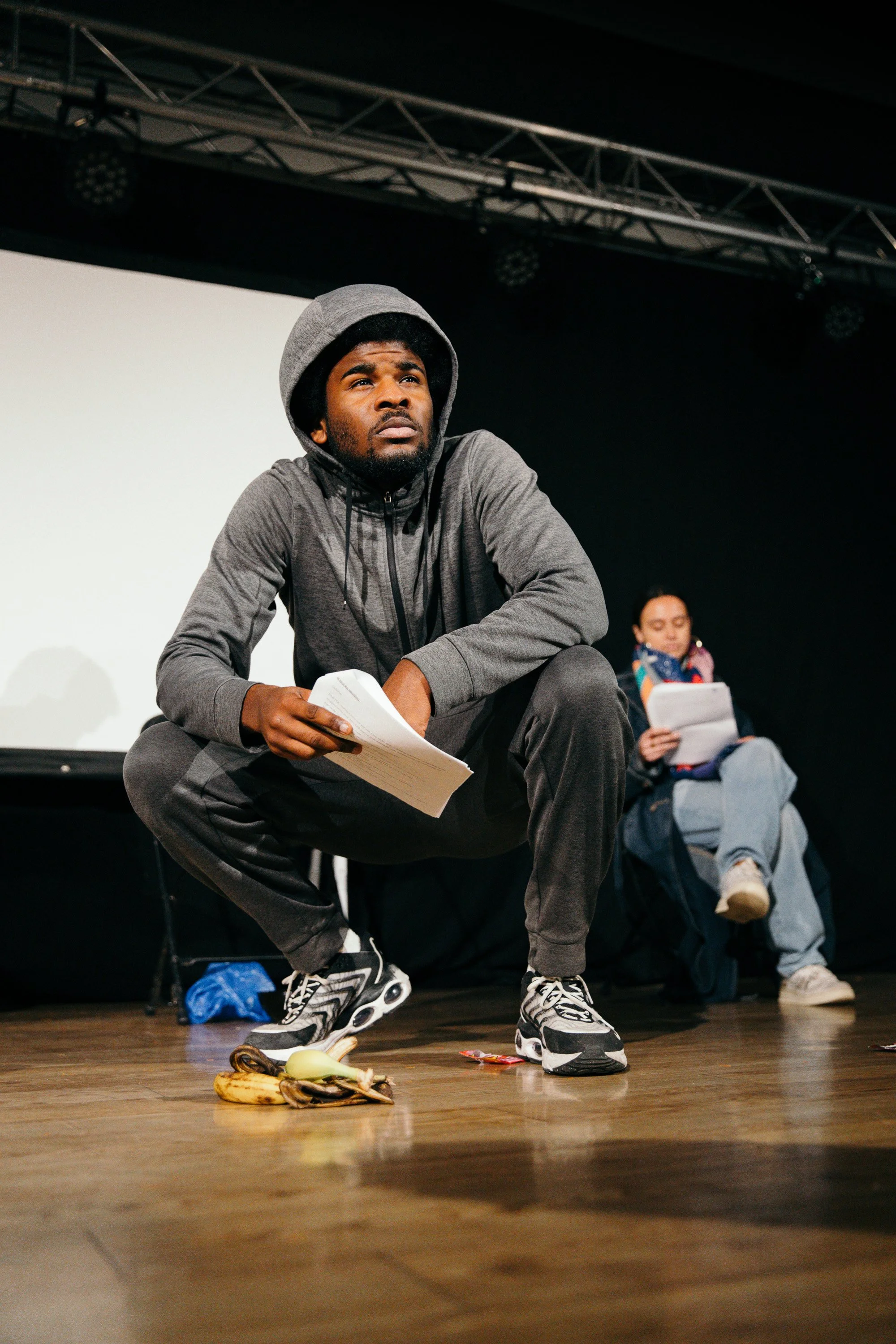 A young man squats on a wooden floor holding papers, with a serious expression, wearing a gray hoodie, gray sweatpants, and athletic shoes. Behind him, a woman sits on a chair reading a script, wearing a jacket, scarf, and jeans. There are bananas, a