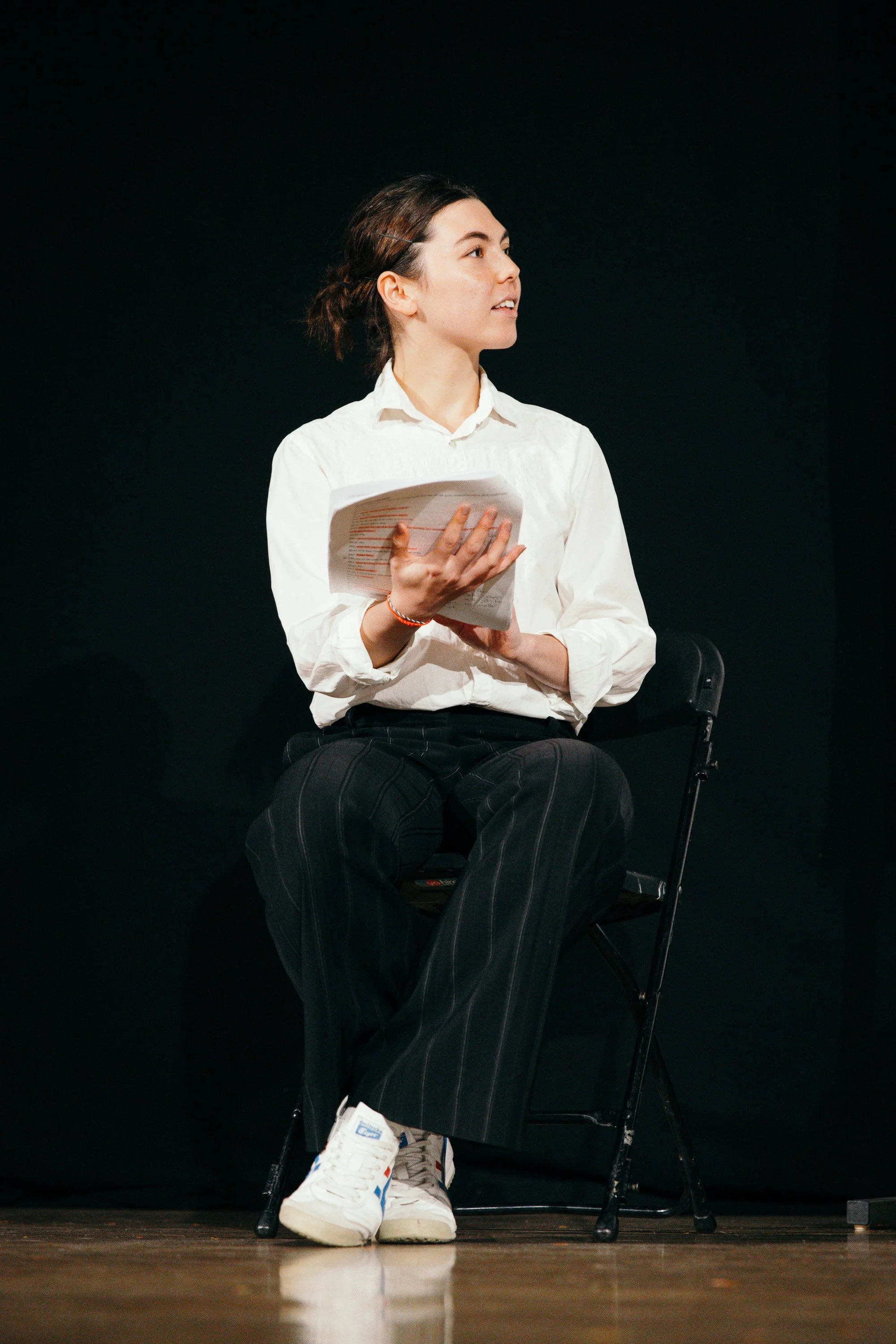 Young woman sitting on a black chair on stage, holding papers, looking to her right, wearing a white shirt, black striped pants, and white sneakers, against a plain black background.