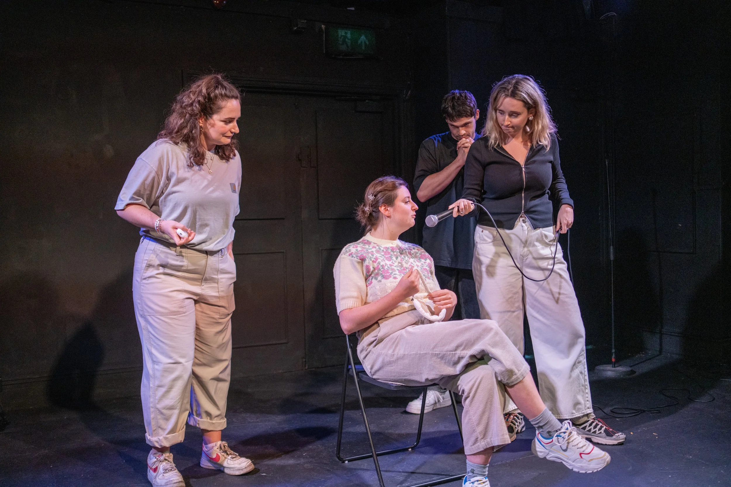 Five people on a stage with dark background, four women and one man, engaged in a theatrical performance. One woman is sitting in a chair, holding a cloth, with a microphone held by another woman standing nearby. The other two women are standing with