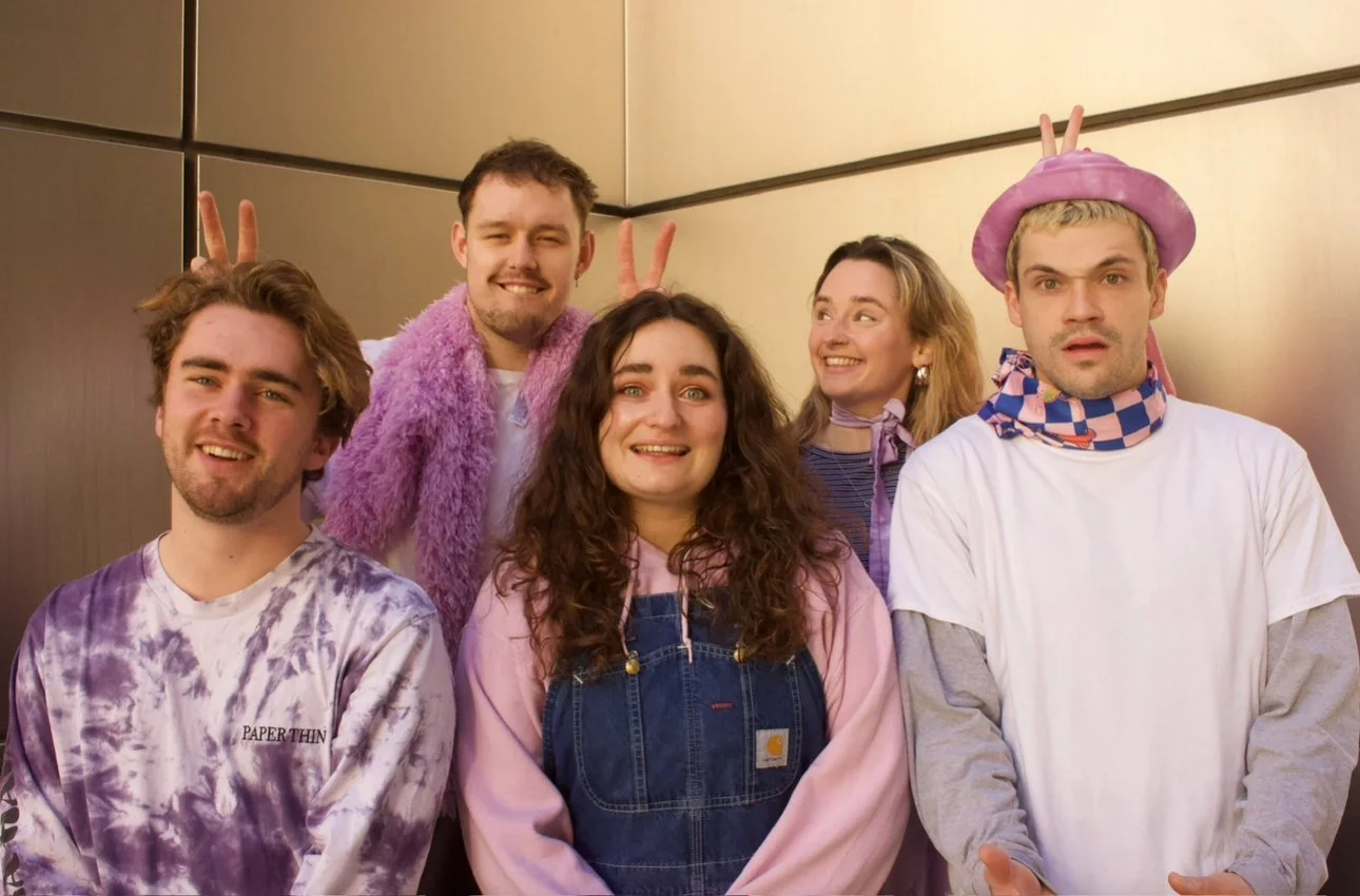 Group of five young adults posing happily for a photo in a metallic elevator. Two of them are making bunny ears behind others.