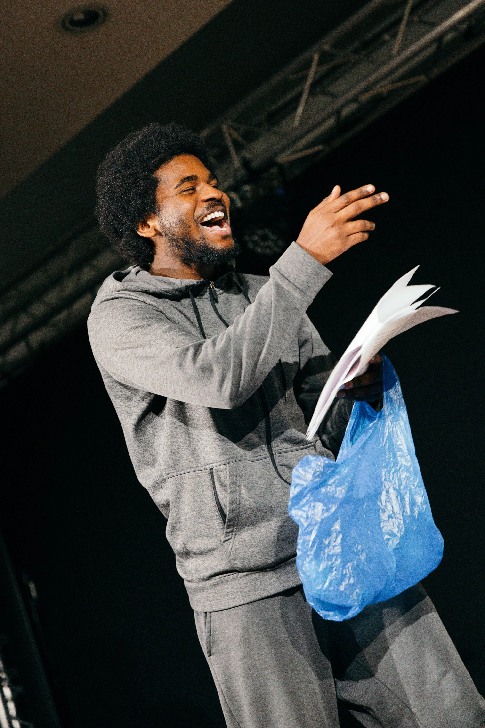 A man with curly hair and a beard, wearing gray hoodie and sweatpants, laughing and gesturing on stage with a clipboard in his hand.