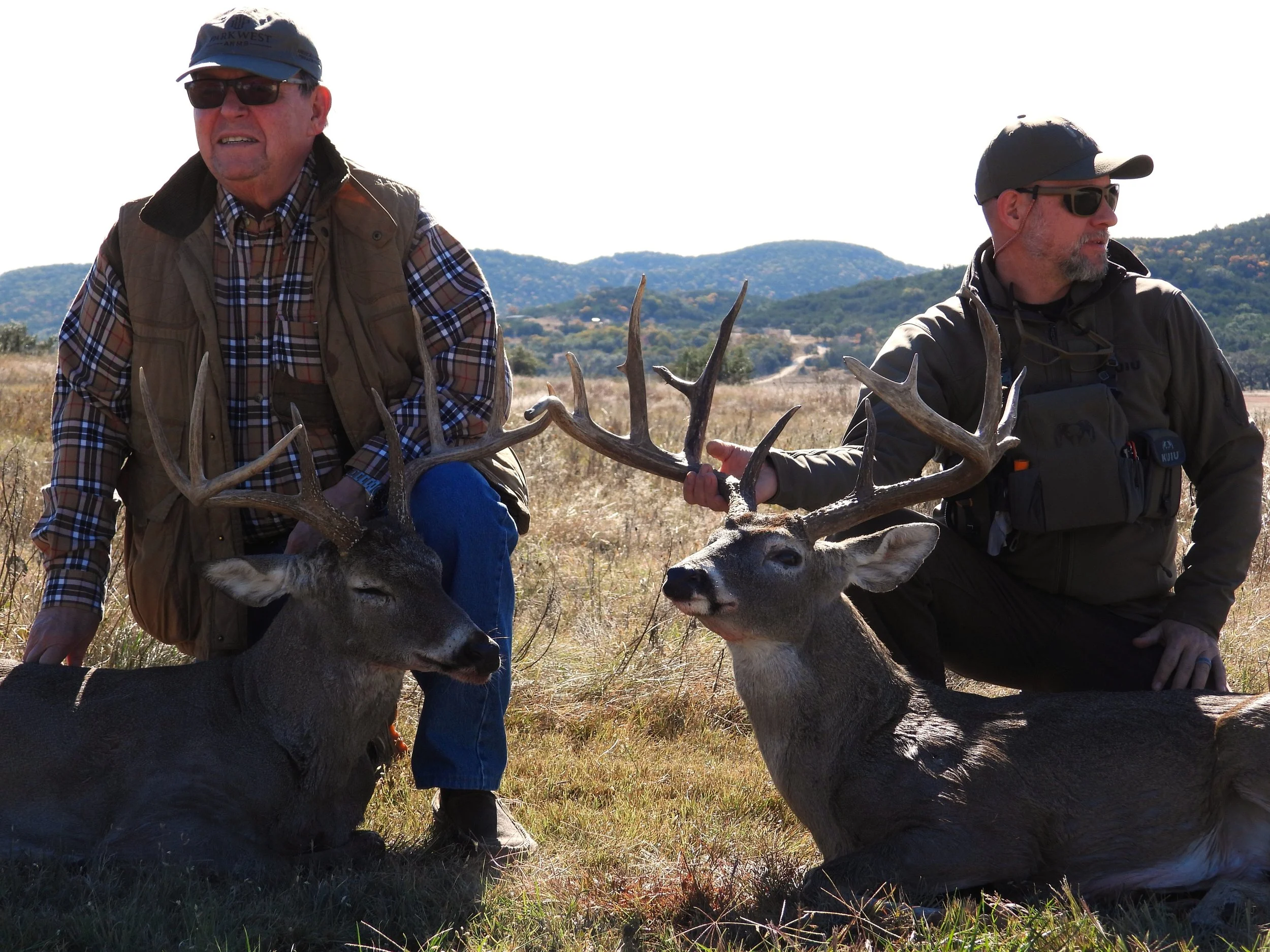 Two men outdoors holding antlers of a deer, with a hilly landscape in the background. The man on the left is wearing a blue cap, sunglasses, and a brown vest over a plaid shirt. The man on the right is wearing a black cap, sunglasses, and a dark jacket.