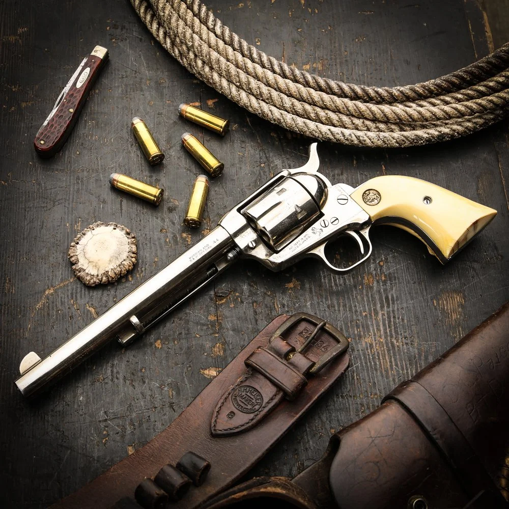 A vintage revolver, a leather gun holster, five bullets, a large knot of rope, a folding pocket knife, a desert plant, and a leather bottle holder are placed on a dark wooden surface.