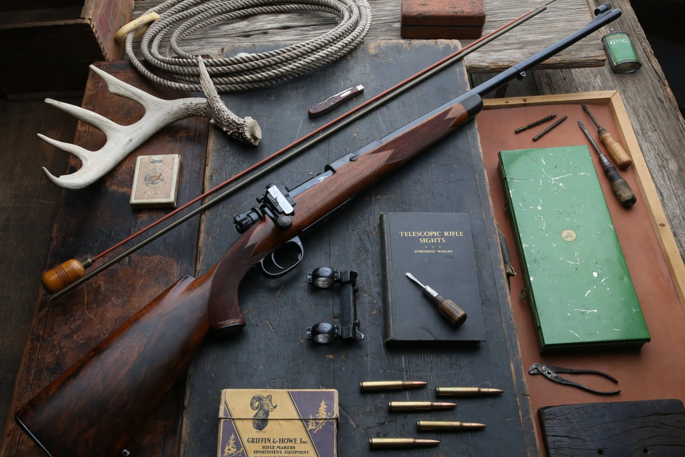 A hunting or shooting setup on a wooden table includes a rifle, bullets, a green box of rifle ammunition, a pair of shooting glasses, a small green book titled "Telescopic Rifle Sights," a large antler, a pocket knife, screwdrivers, and other small tools, along with a coil of rope and some boxes.