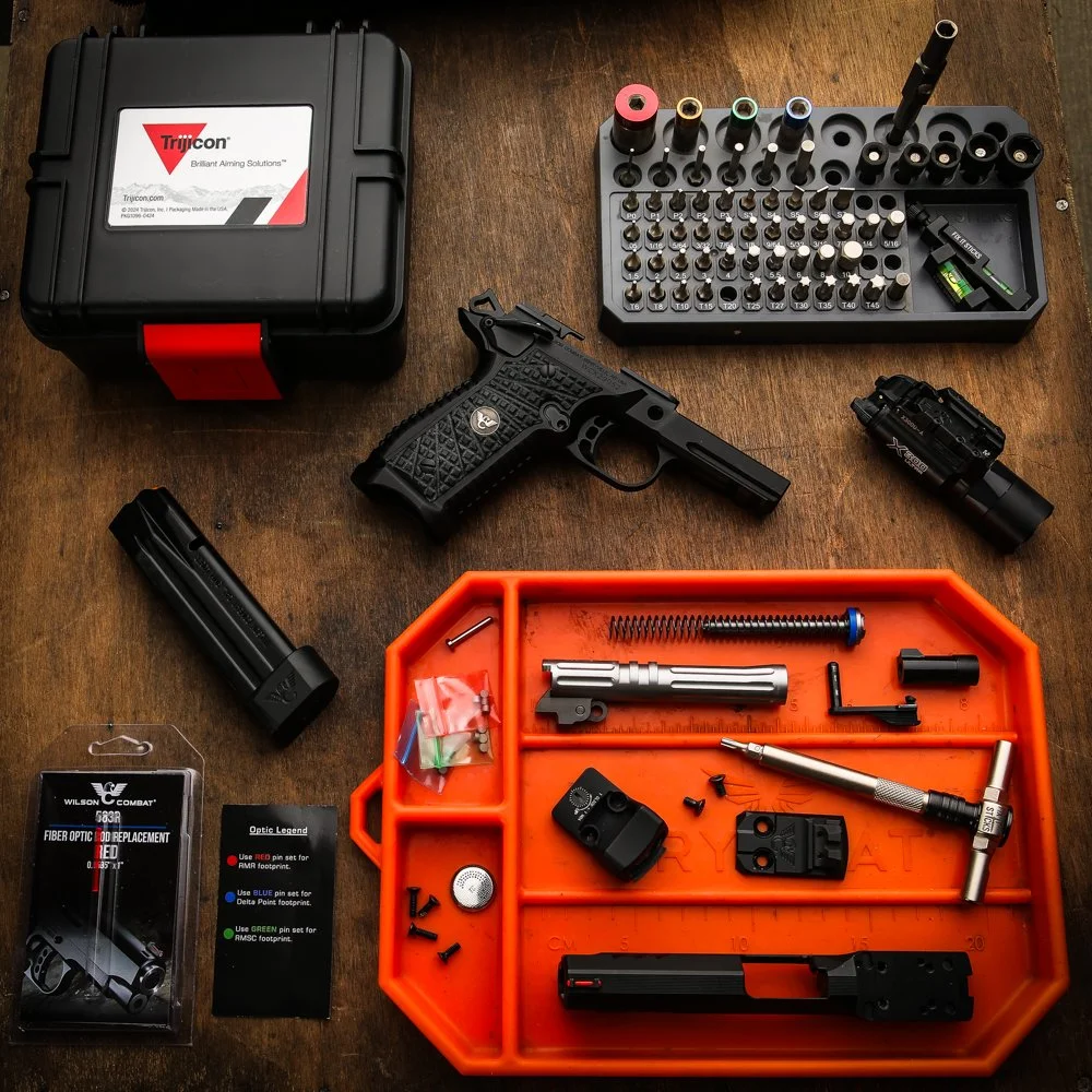 Gun parts, tools, and accessories laid out on a wooden surface, including a black pistol, a magazine, a scope, a cleaning kit, and various small parts and screws.