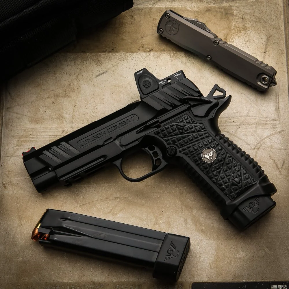 A black Wilson Combat handgun with a textured grip, a detached magazine, and a standalone upper slide placed on a wooden surface.