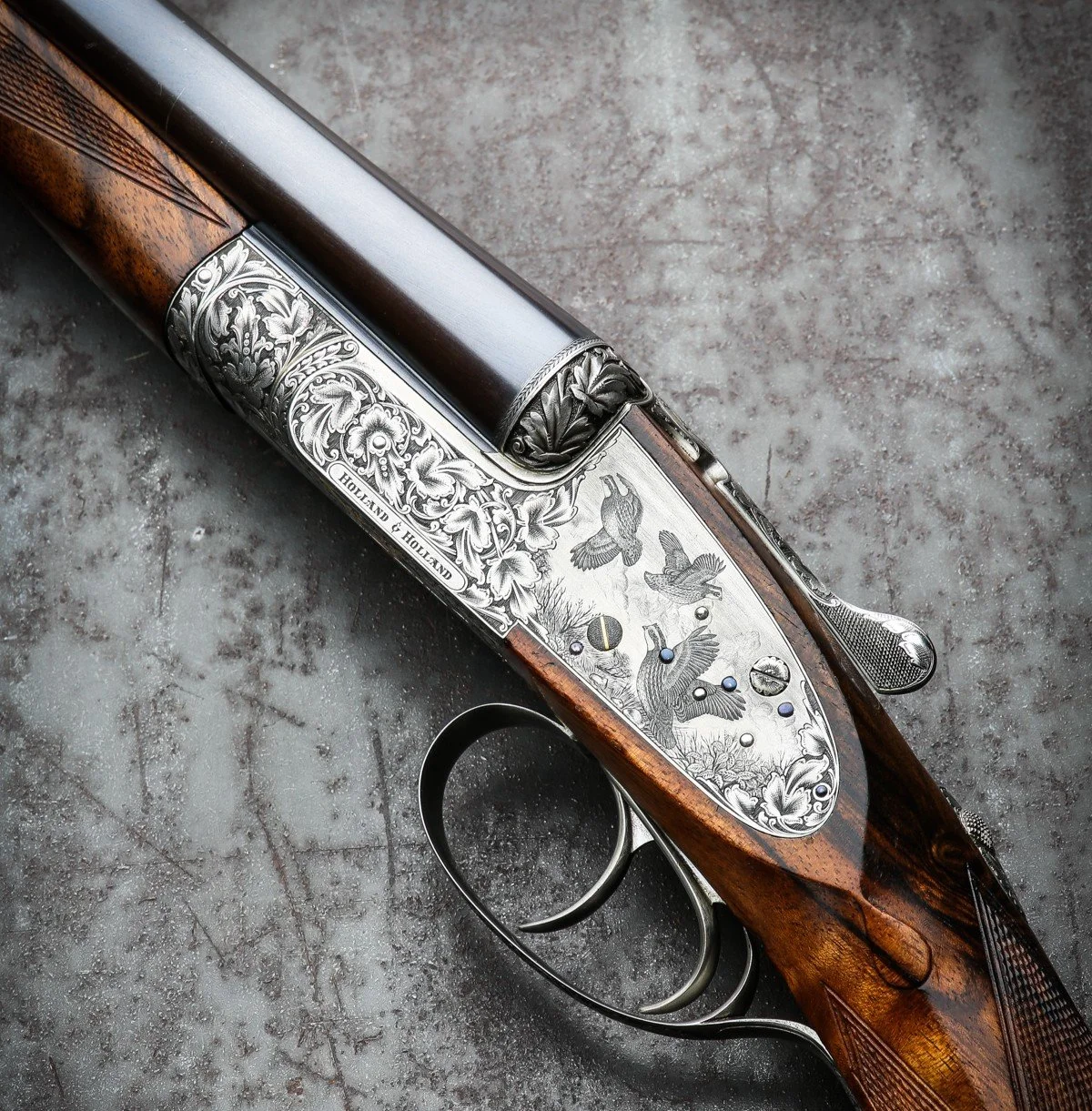A detailed hunting rifle with intricate silver engraving on the receiver, featuring birds and floral patterns, resting on a textured surface.