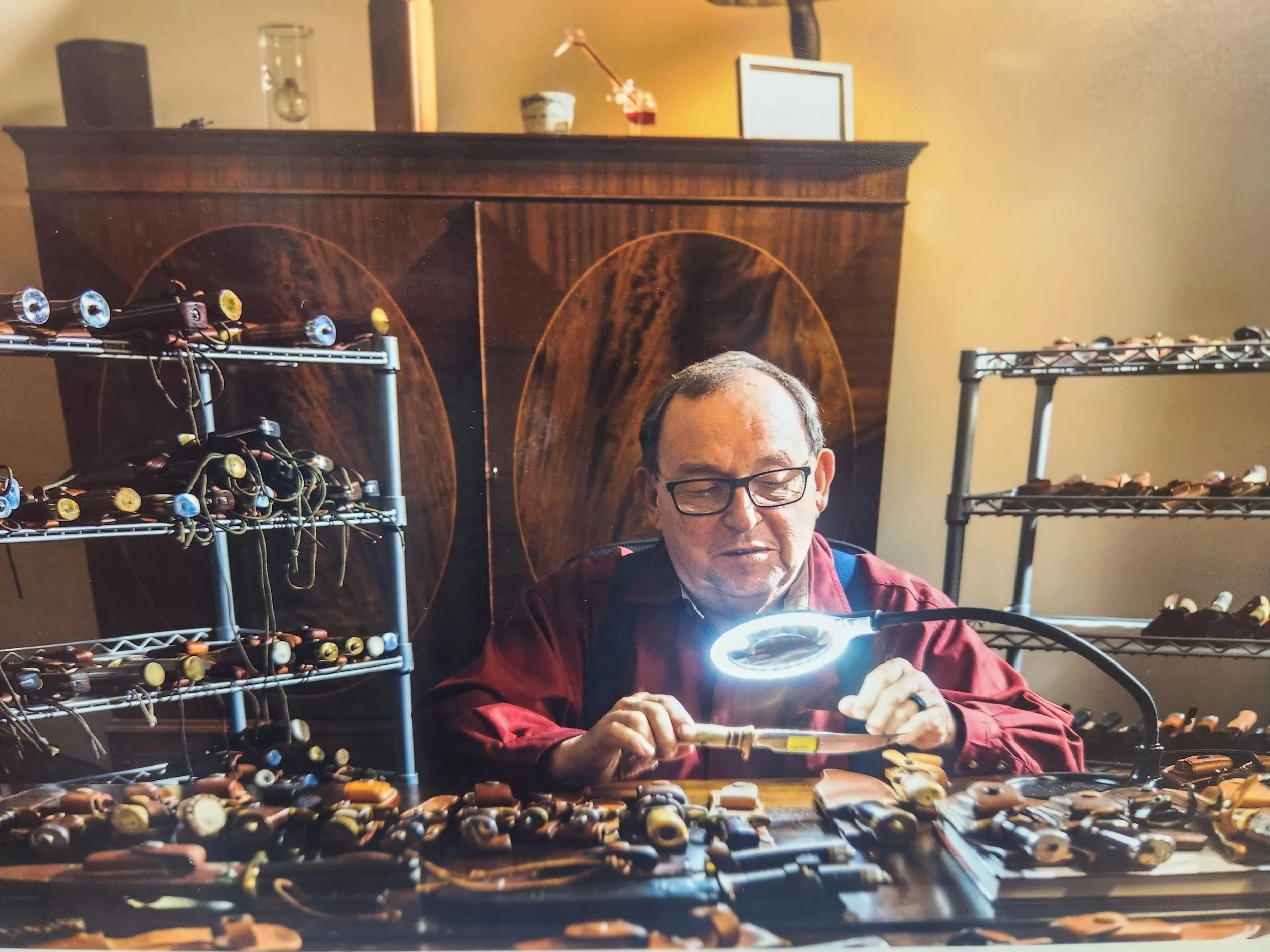 A man with glasses and a red jacket is working on a rifle at a table filled with rifle parts and components, with metal shelves holding more rifle parts in the background.
