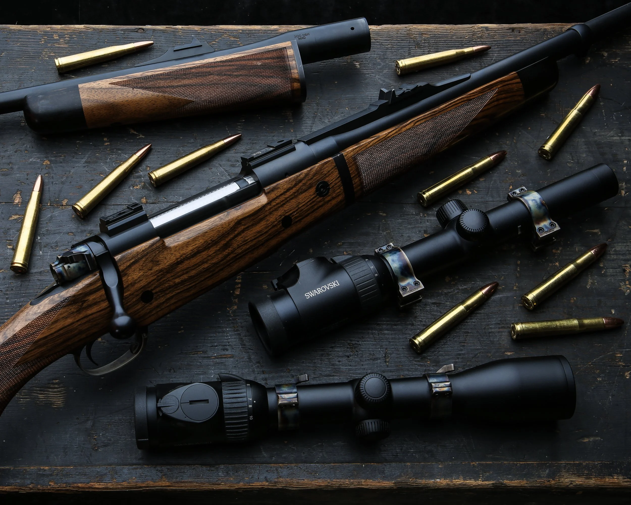 A pair of rifles with wooden stocks lying on a dark wooden surface, surrounded by several rounds of ammunition and a scope attached to one of the rifles.