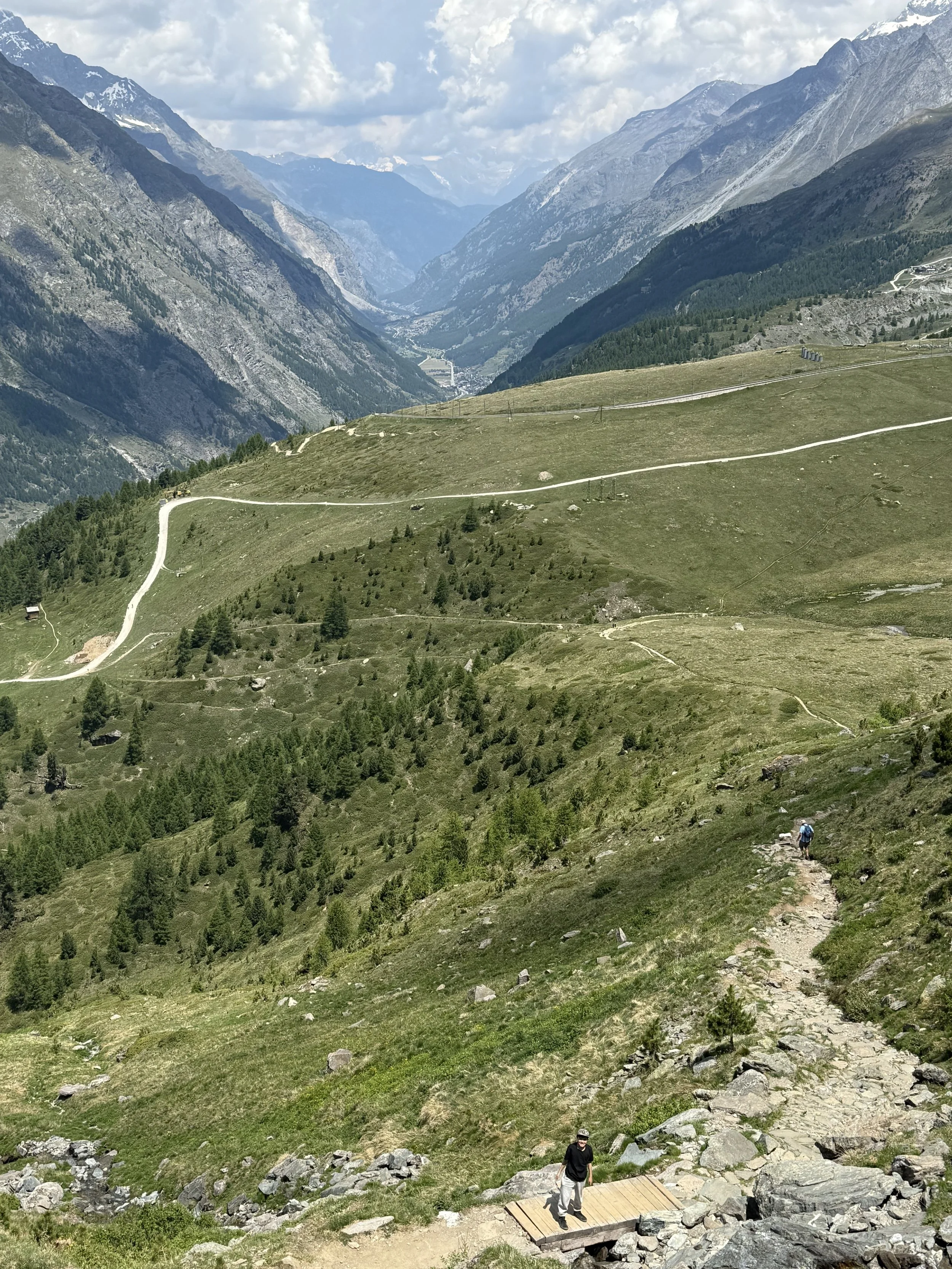 Hiking trail through a lush green mountain landscape with hikers walking along the path, surrounded by mountain ranges and a partly cloudy sky.  Journey through immigration.