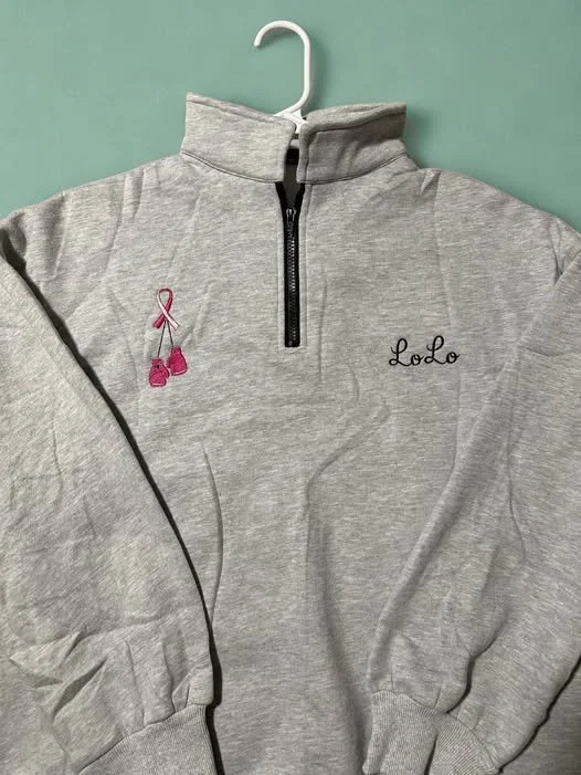 Gray quarter-zip sweatshirt with embroidered pink boxing gloves and a name "LoLo" on the front, hanging on a white hanger against a green wall.