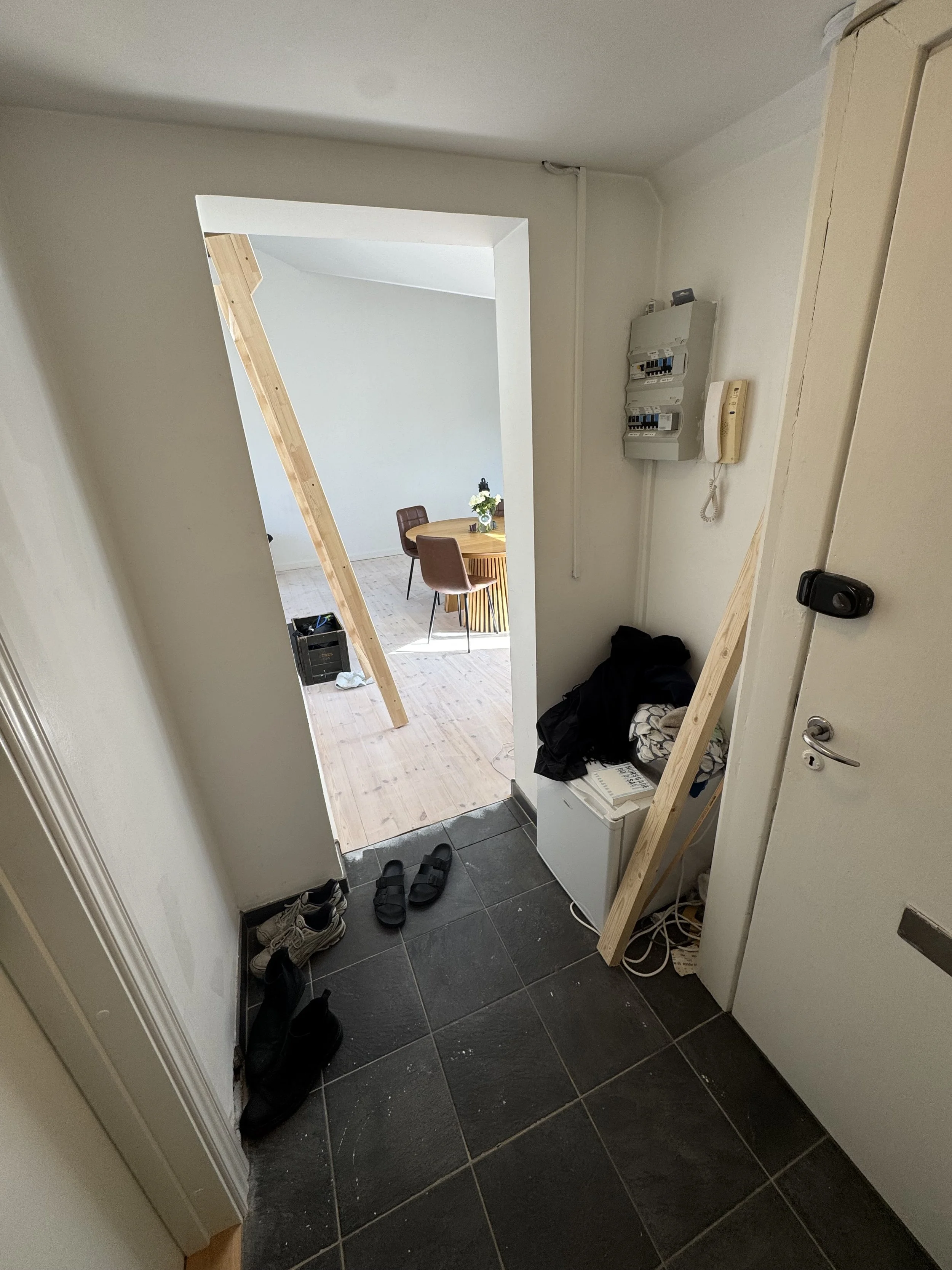 Entryway with black tiled floor, shoes and sandals on the floor, wooden beams leaning against the wall, an open door showing the living room with wood flooring, a small round dining table with chairs, and a window with sunlight.