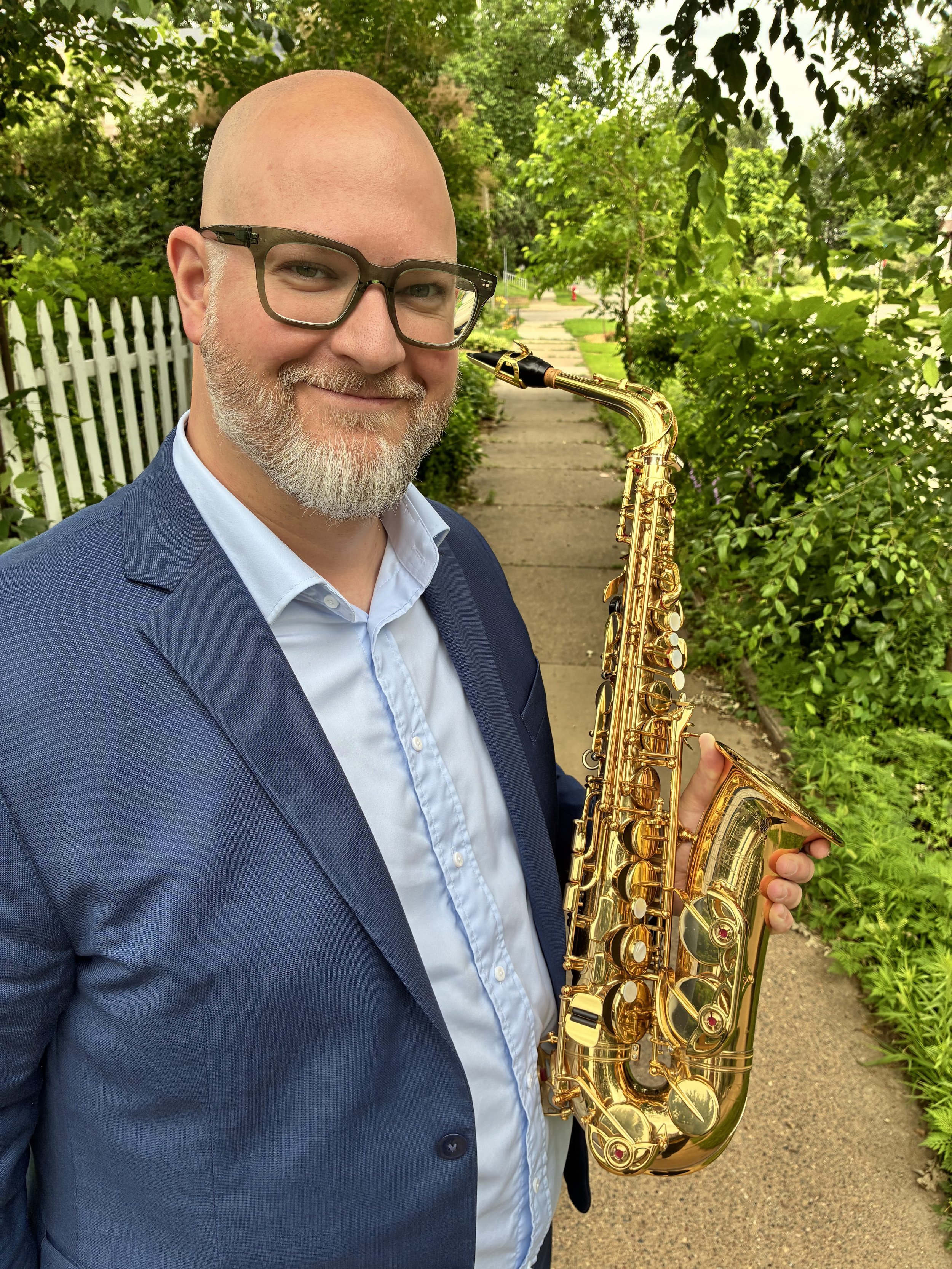 A man with glasses, a beard, and a mustache wearing a blue suit jacket and light blue shirt, smiling while holding a gold saxophone outdoors on a sidewalk with green trees and bushes in the background.