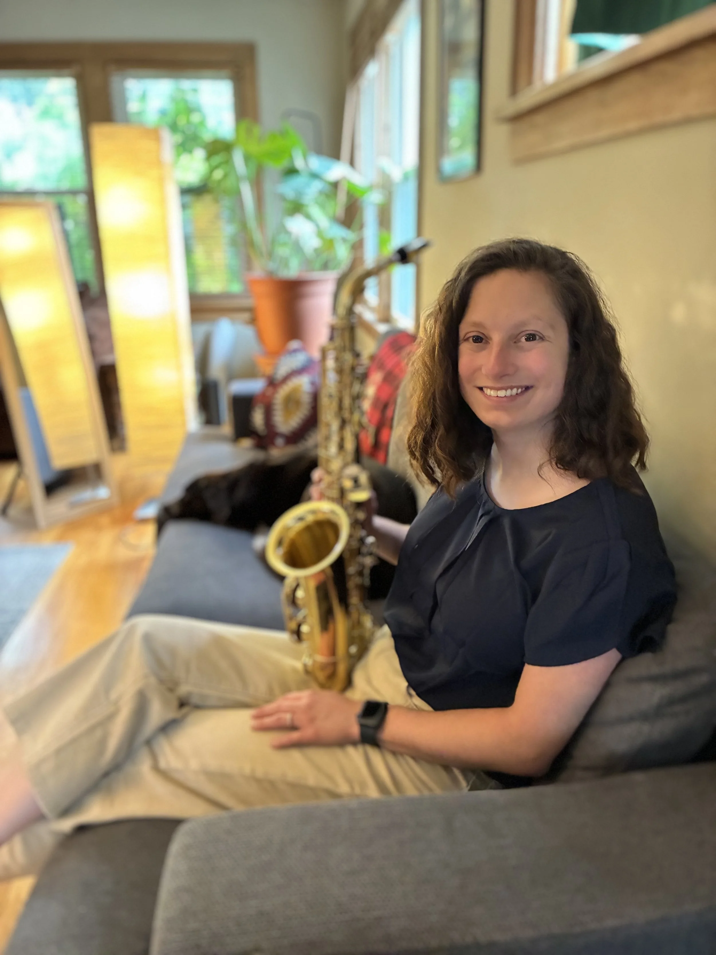 A woman with curly brown hair sitting on a gray couch holding a gold-colored saxophone, smiling at the camera inside a cozy, sunlit living room.