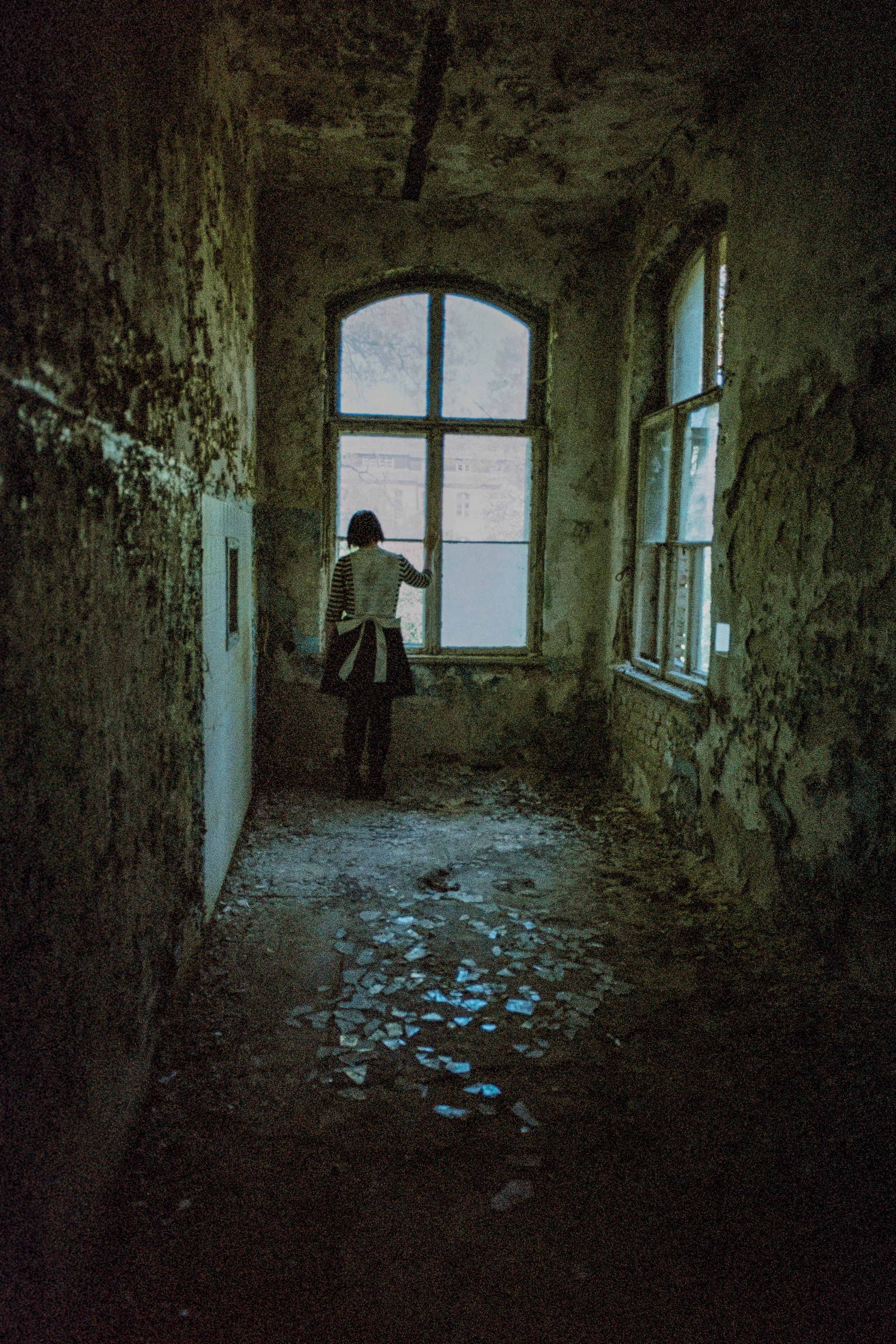 Eine Person schaut durch ein altes, zerfallenes Fenster in einem verfallenen Gebäude mit beschmutzten Wänden und zerbrochenem Boden.