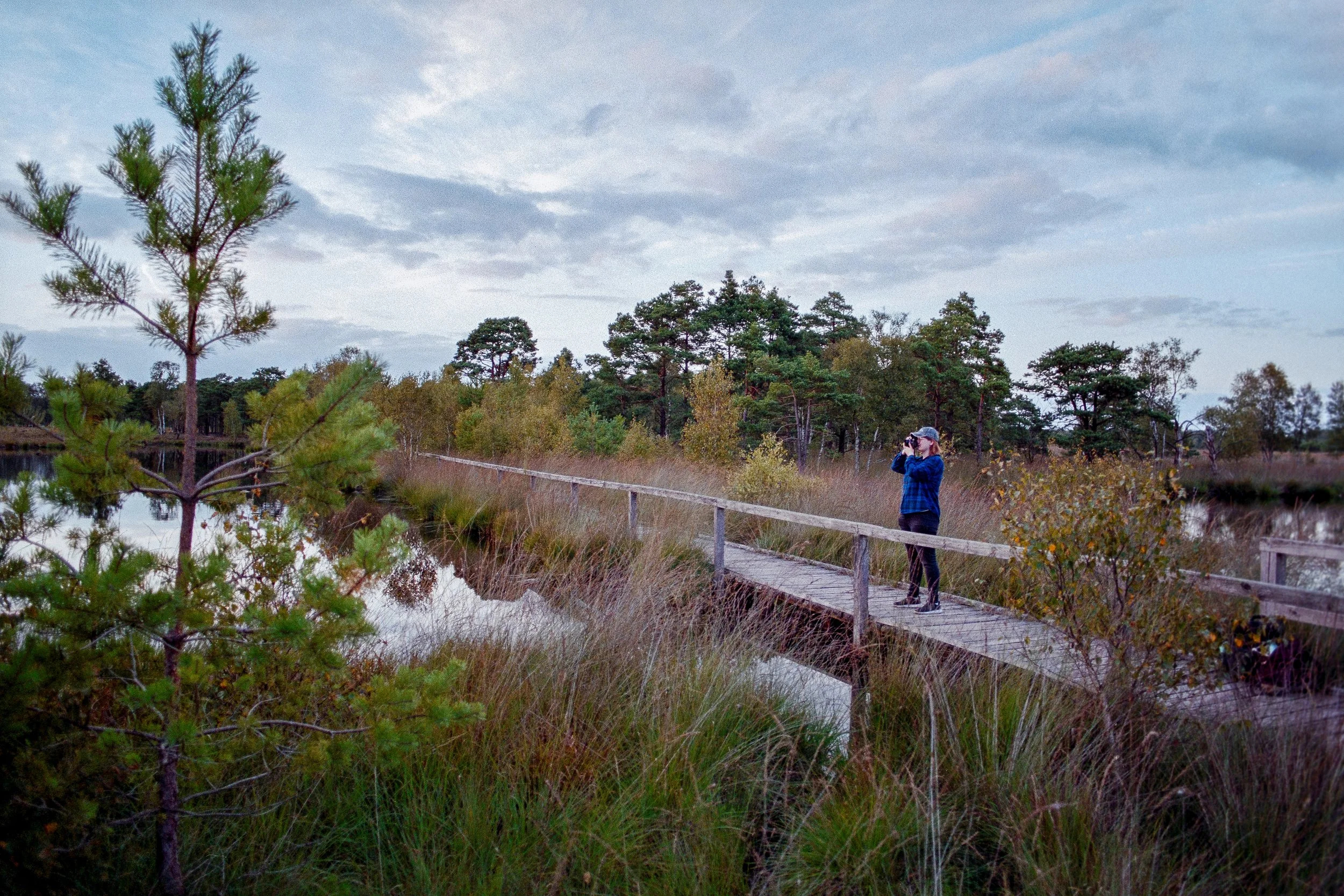 Person steht auf einem Holzsteg in einer natürlichen Landschaft und fotografiert mit einer Kamera, umgeben von Bäumen und Wasser, Himmel mit Wolken