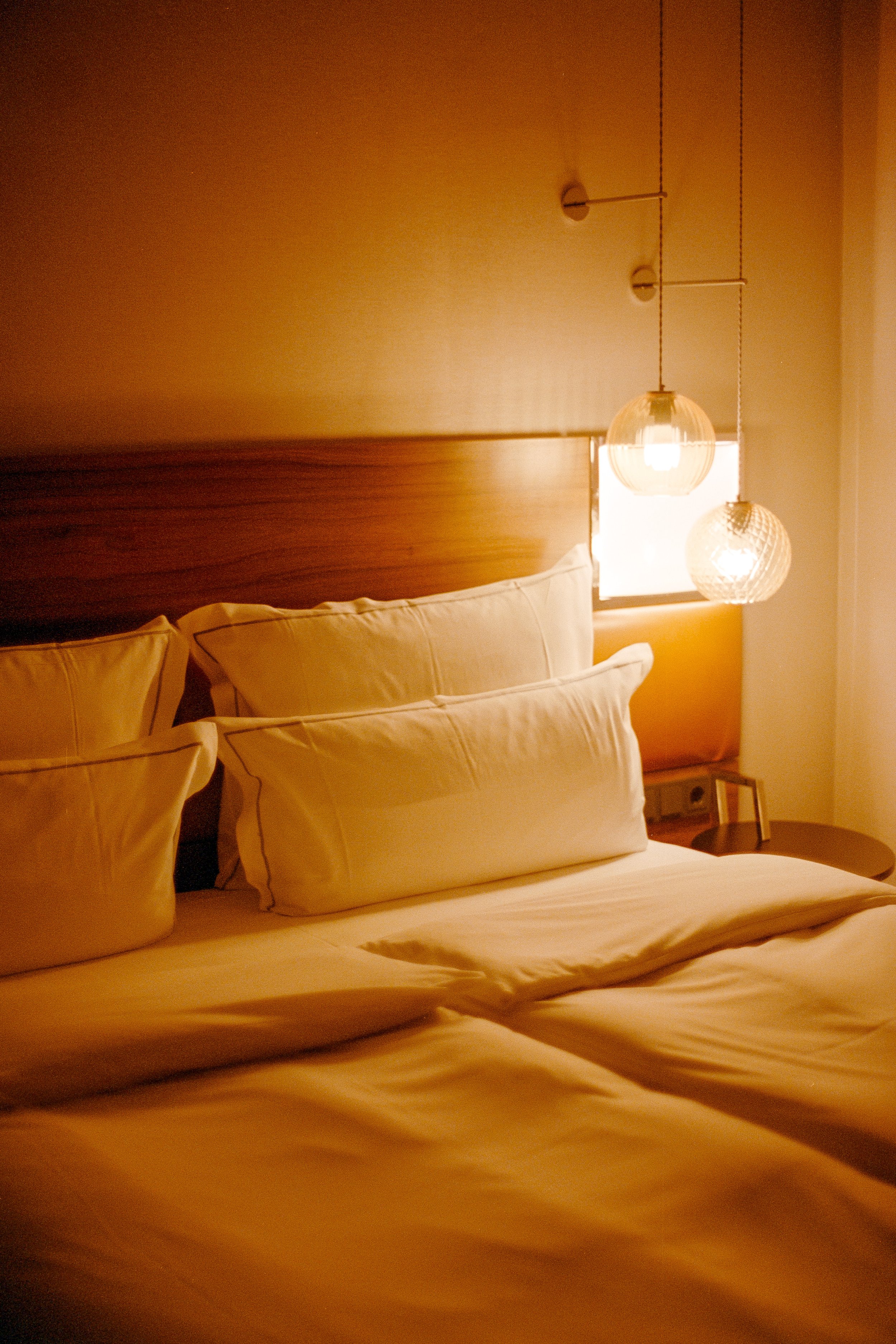 Ein gemütliches Schlafzimmer mit einem Bett, Bettwäsche und Kissen, neben einem Fenster mit zwei hängenden Lamellenleuchten und warmem Licht.