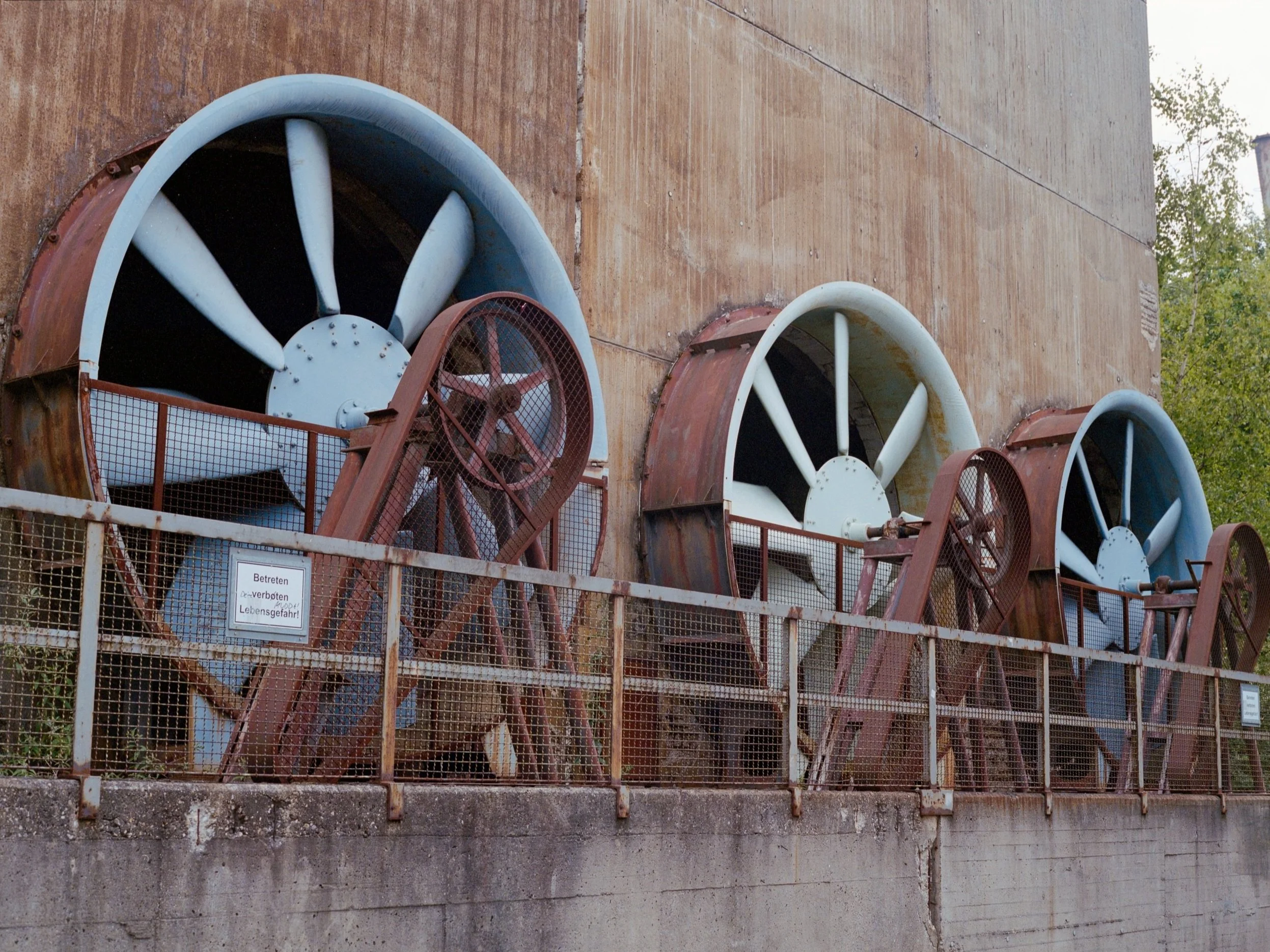 Alte Industriewindturbinen an einer Wand, umgeben von einem Metallzaun mit Warnschild.