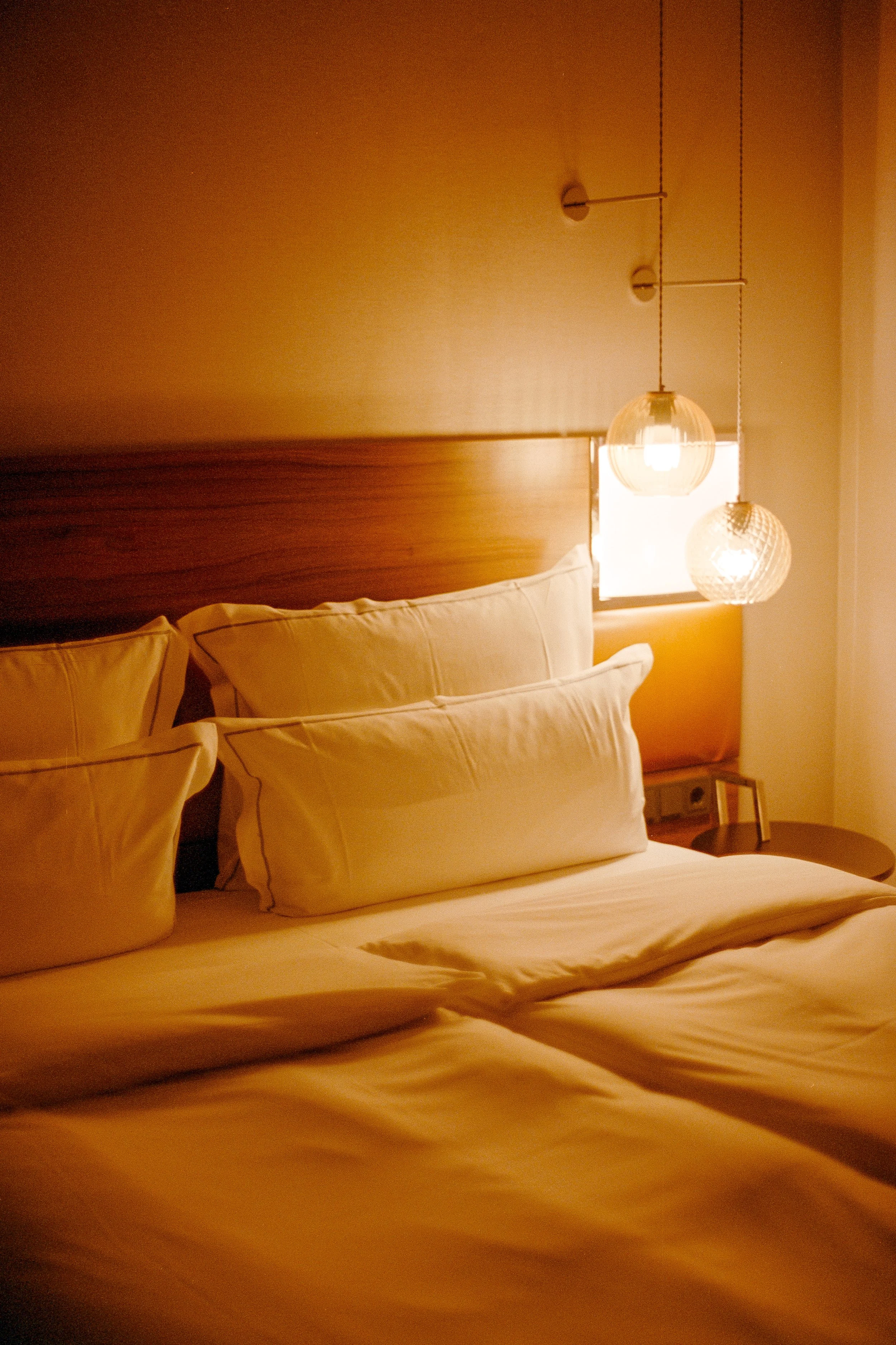 Gemütliches Schlafzimmer mit Bett, weißen Kissen, Holzwandkopfteil und two hängenden Lampen, in warmem Licht.