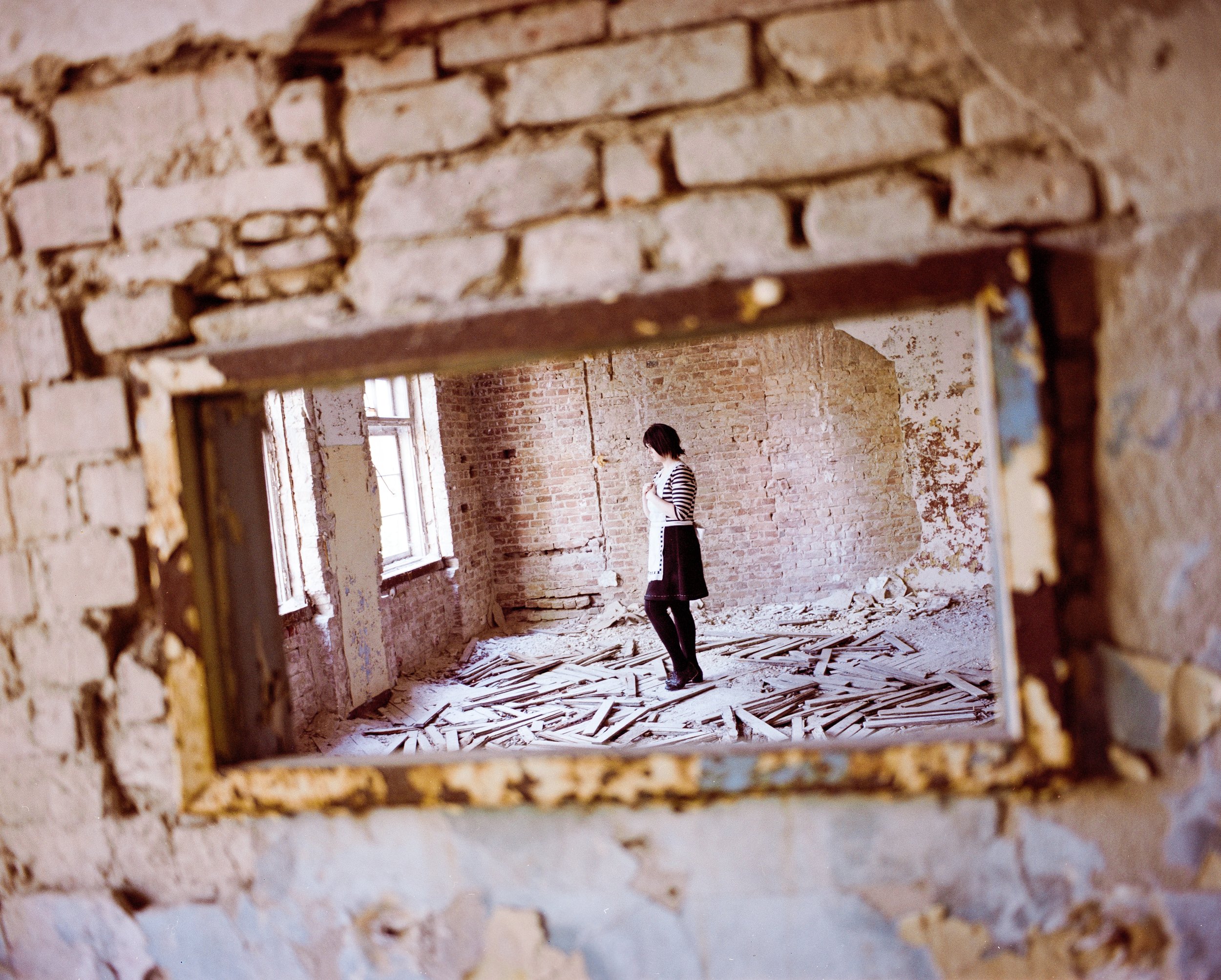 Frau steht in einem unfertigen, alten Backsteinhaus mit zerbrochenen Dielen auf dem Boden, Blick durch eine kleine Maueröffnung.