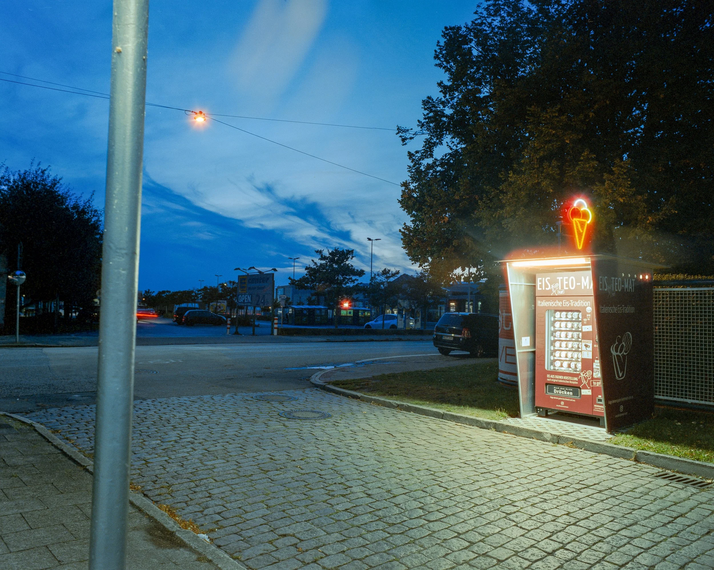 Eisautomat mit Neon-Schild in einer Stadt bei Dämmerung, umgeben von Bäumen und Autos.