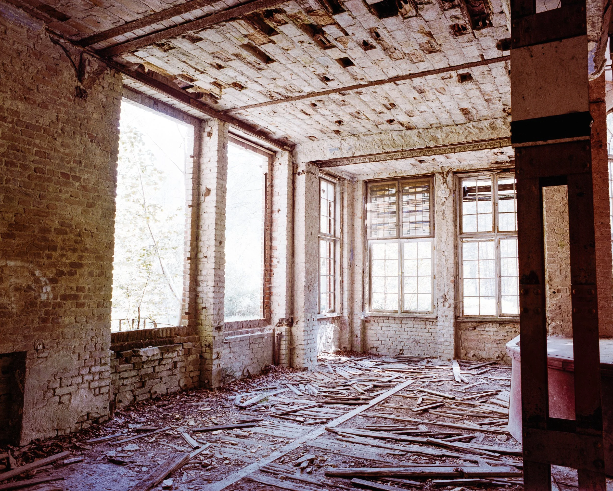 Innenansicht eines verlassenen, verfallenen Gebäudes mit bröckelnden Wänden, großen Fenstern und einem Boden voller Holz- und Baumaterialreste.