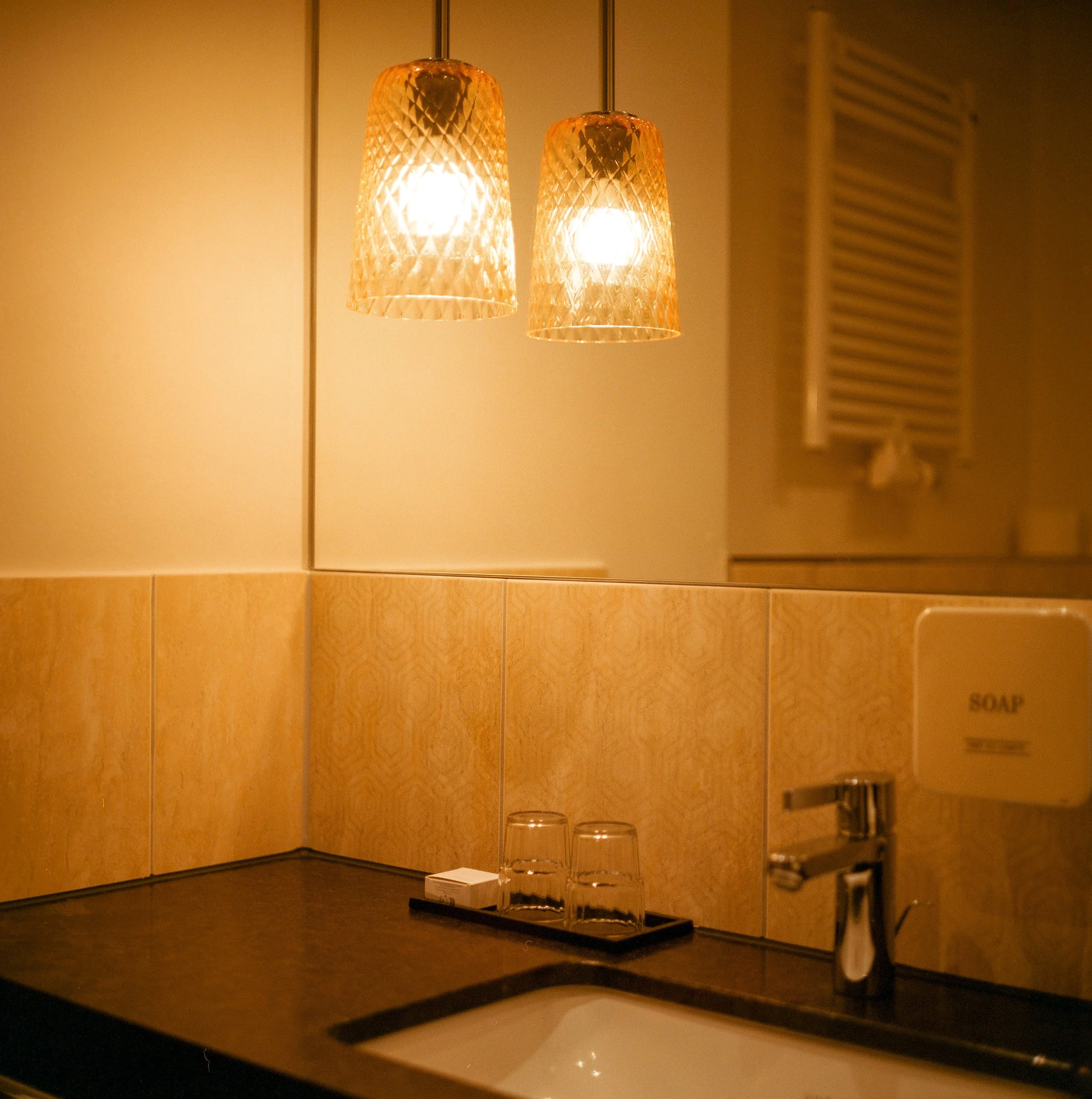 Waschbecken mit zwei Trinkgläsern, Seifenhalter mit der Aufschrift 'SOAP', Hängelampen, Wandspiegel, dunkler Waschbeckenbereich, lichtdurchflutender Raum