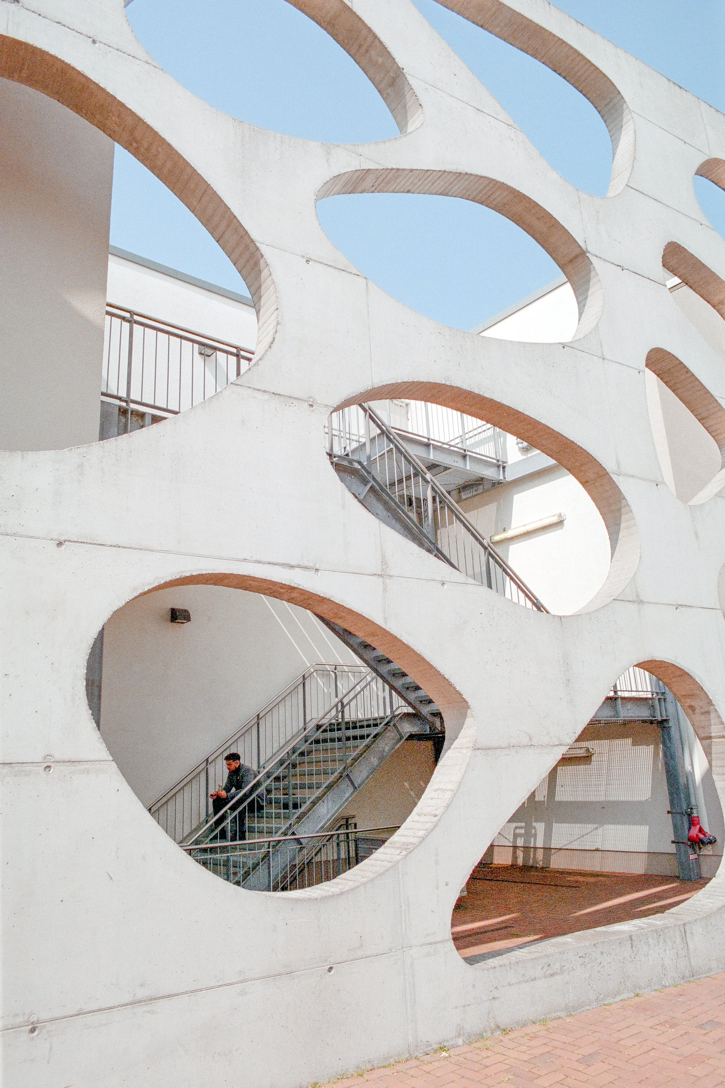 Ein moderner Gebäudeteil mit großen ovale Öffnungen und einem Außenaufstieg, durch den eine Person die Treppe hinaufgeht.