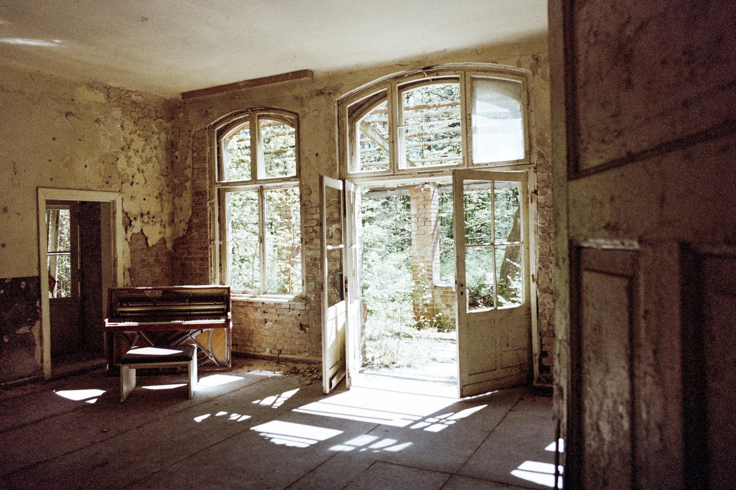 Verlassener Raum mit alten, zerbröckelnden Wänden und großen Fenstern durch die Tageslicht fällt. Es befindet sich ein alter Klavierstuhl und ein beschädigter Klavier im Raum.