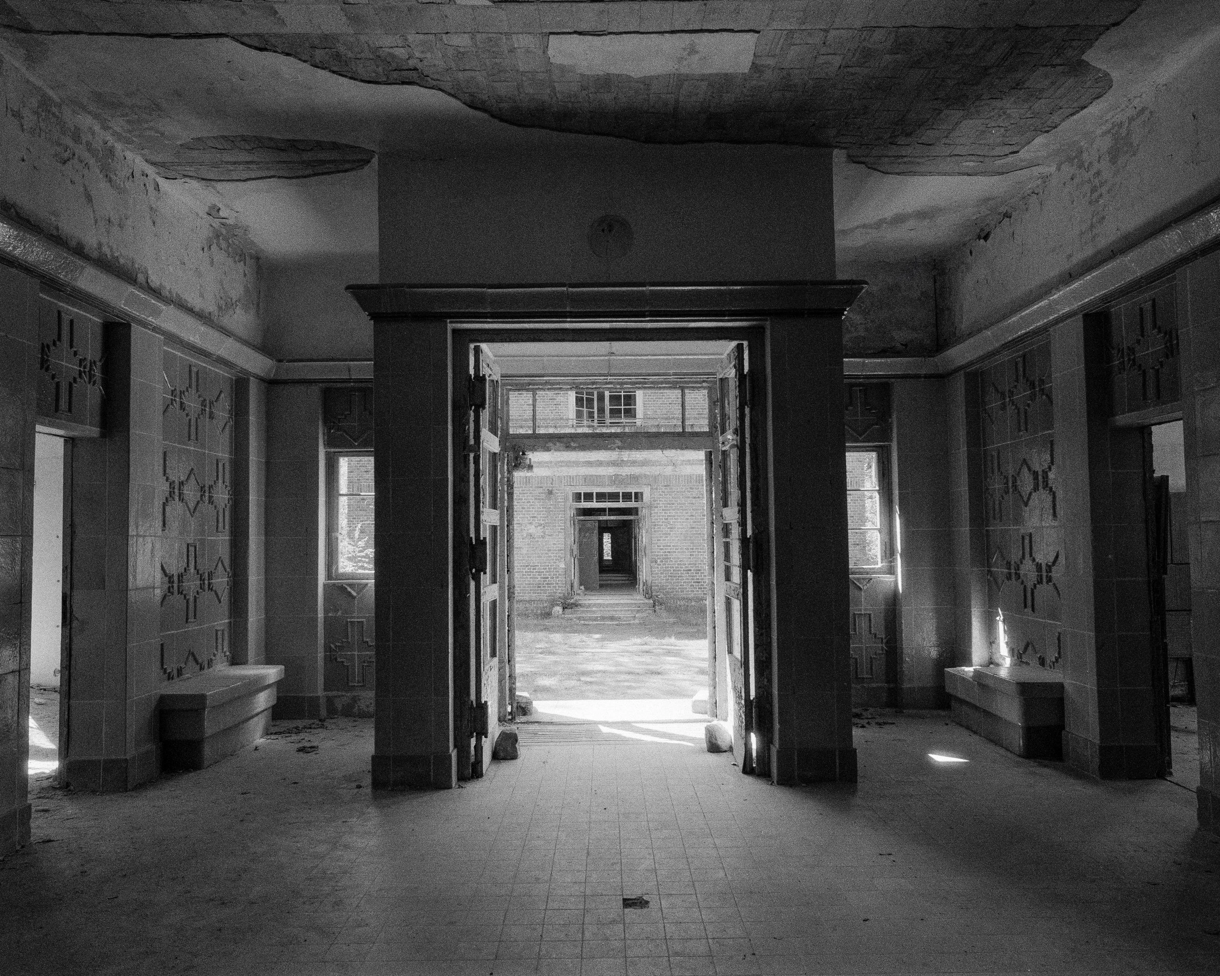 Ein verlassener, leerer Raum in einem Gebäude mit offenen Türen, durch die man auf einen Hof mit weiteren Gebäuden blickt, die Fenster sind zerbrochen und Wände abgenutzt.